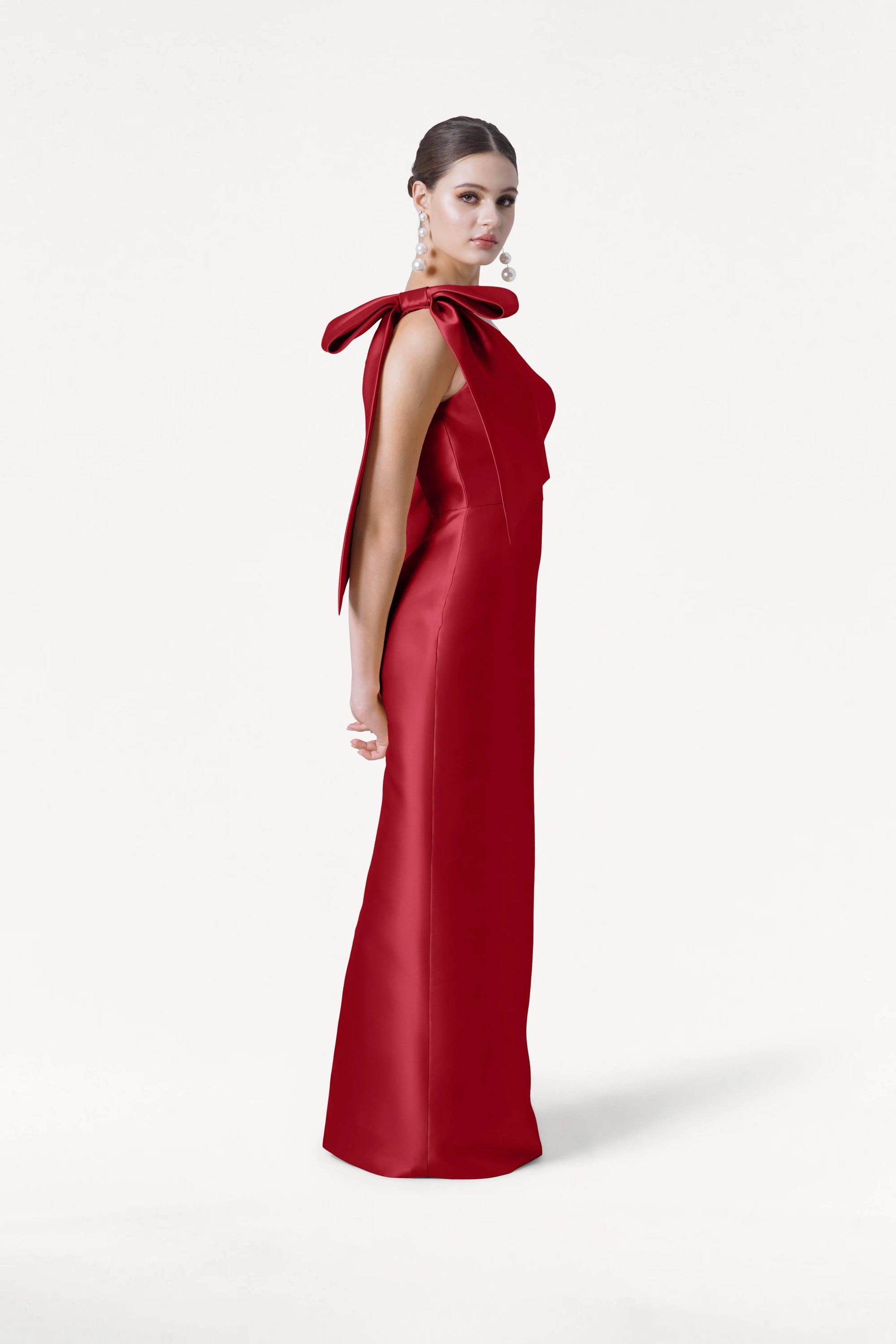 A woman in the TH&TH Ritz Bridesmaid Dress in Dark Cherry Satin Twill, featuring large shoulder bows, stands against a white background, looking over her shoulder with her hair styled in a neat bun.