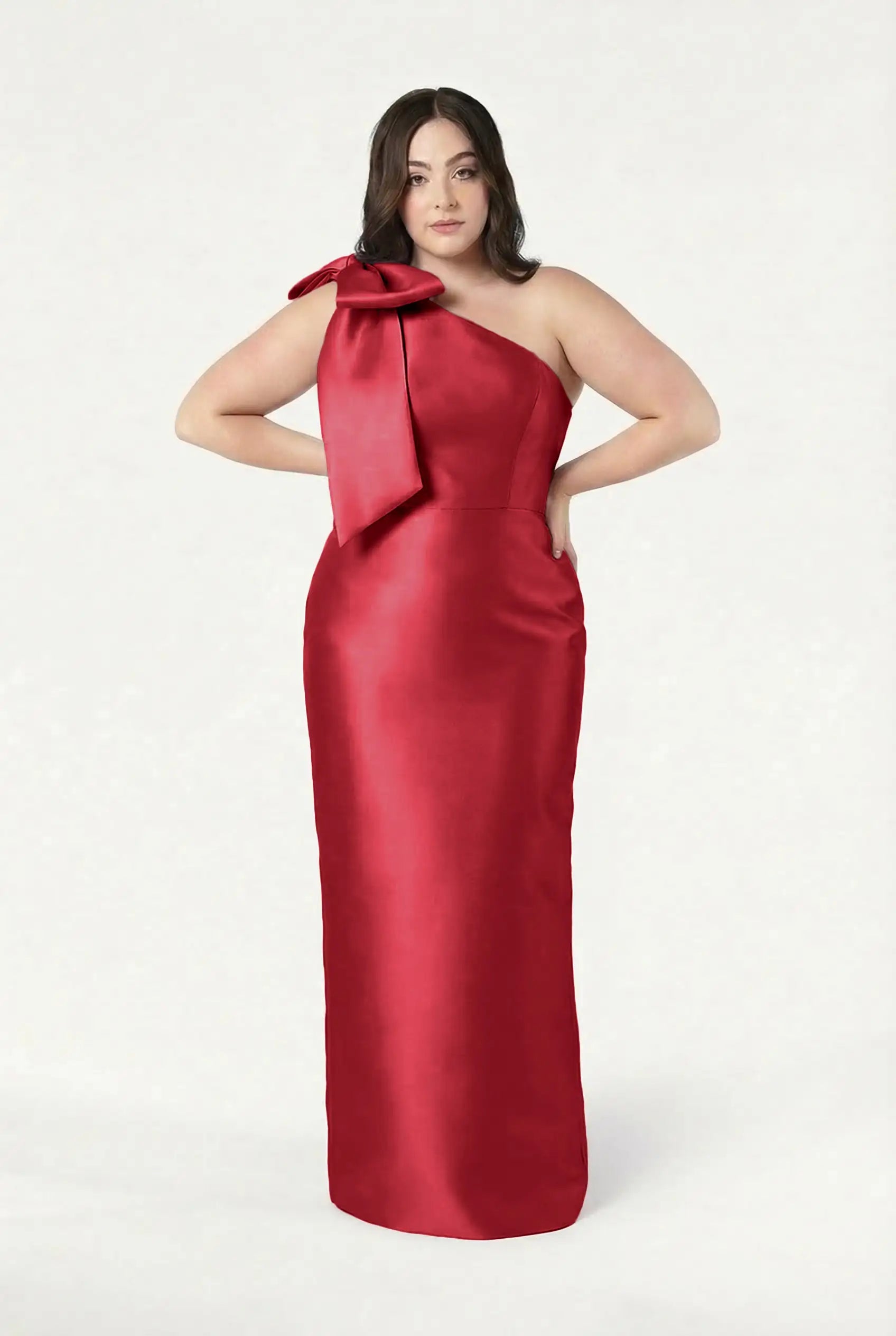 A woman wearing the TH&TH Ritz Bridesmaid Dress in Dark Cherry Satin Twill, featuring a one-shoulder neckline and large bow, stands against a plain white background with her hands casually in her pockets.