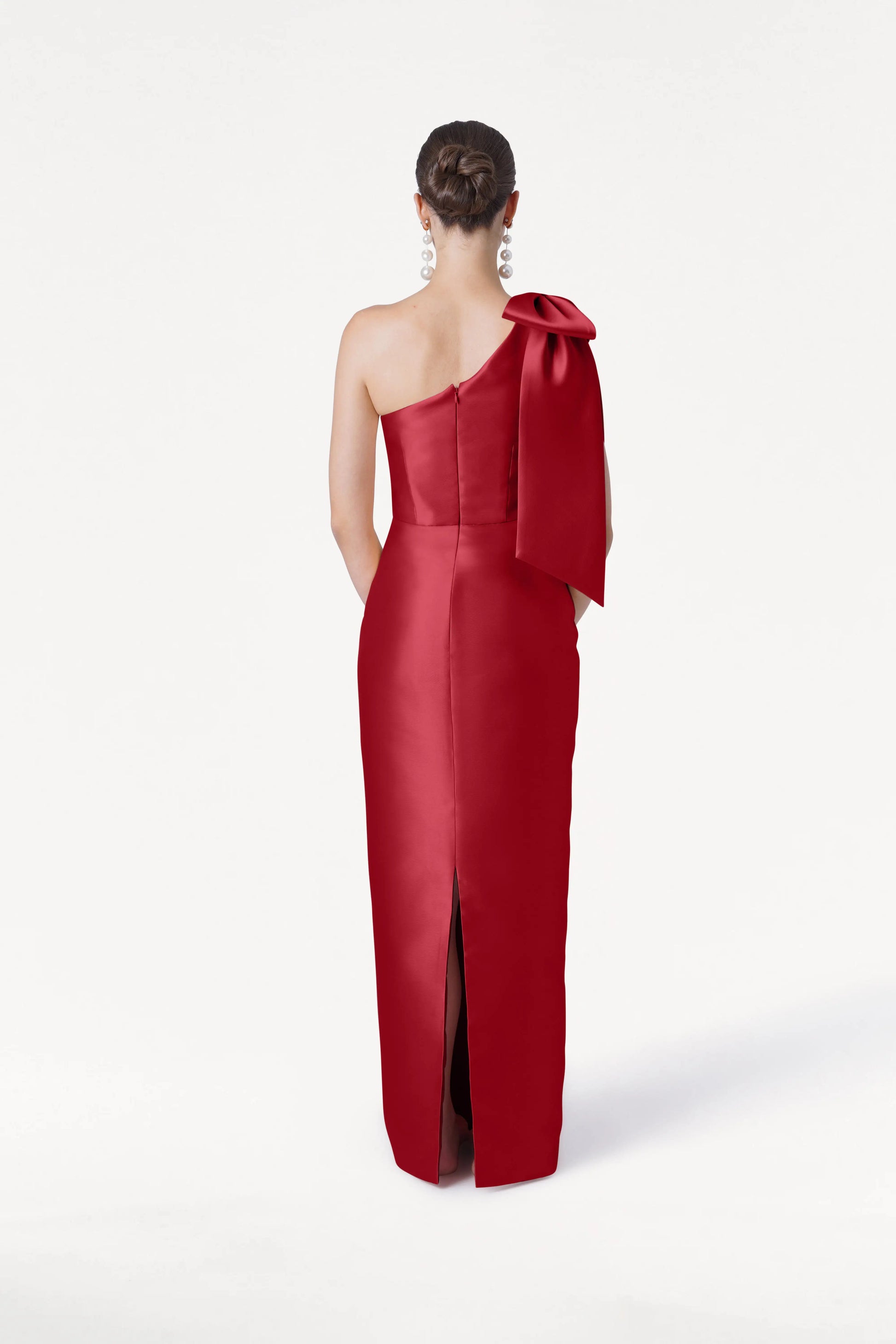 A woman stands with her back to the camera in the TH&TH Ritz Bridesmaid Dress in Dark Cherry Satin Twill, a floor-length, one-shoulder gown featuring a large bow on the shoulder. Her hair is in a neat bun and she wears statement earrings.