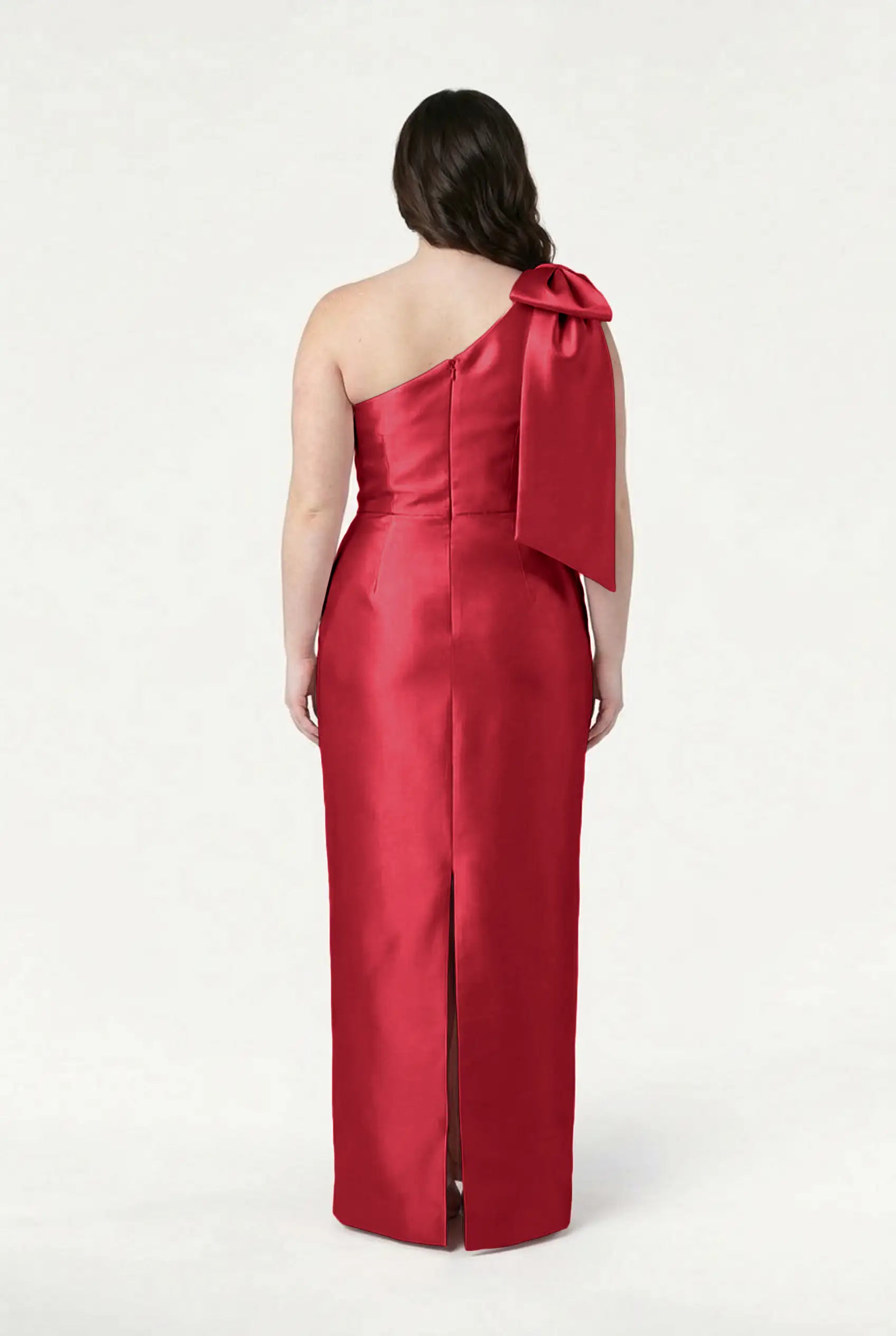 A woman faces away in the TH&TH Ritz Bridesmaid Dress in Dark Cherry Satin Twill, a long, fitted one-shoulder gown with a large bow on the left shoulder and a center back slit. The background is plain white.