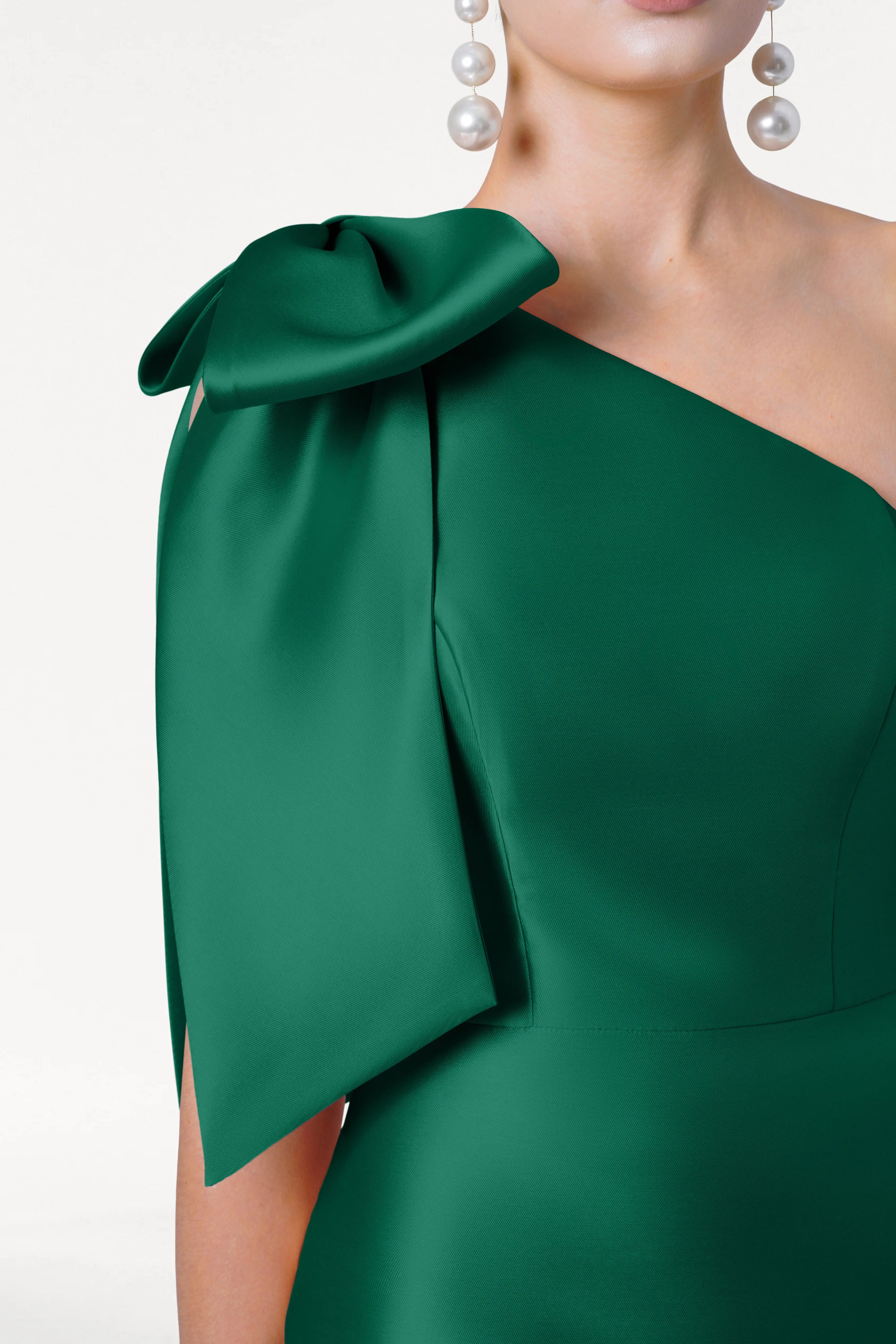 A woman wears the TH&TH Ritz Bridesmaid Dress in Forest Green Satin Twill, featuring a one-shoulder design with a statement bow and pearl drop earrings, shown from shoulders to waist against a plain background.