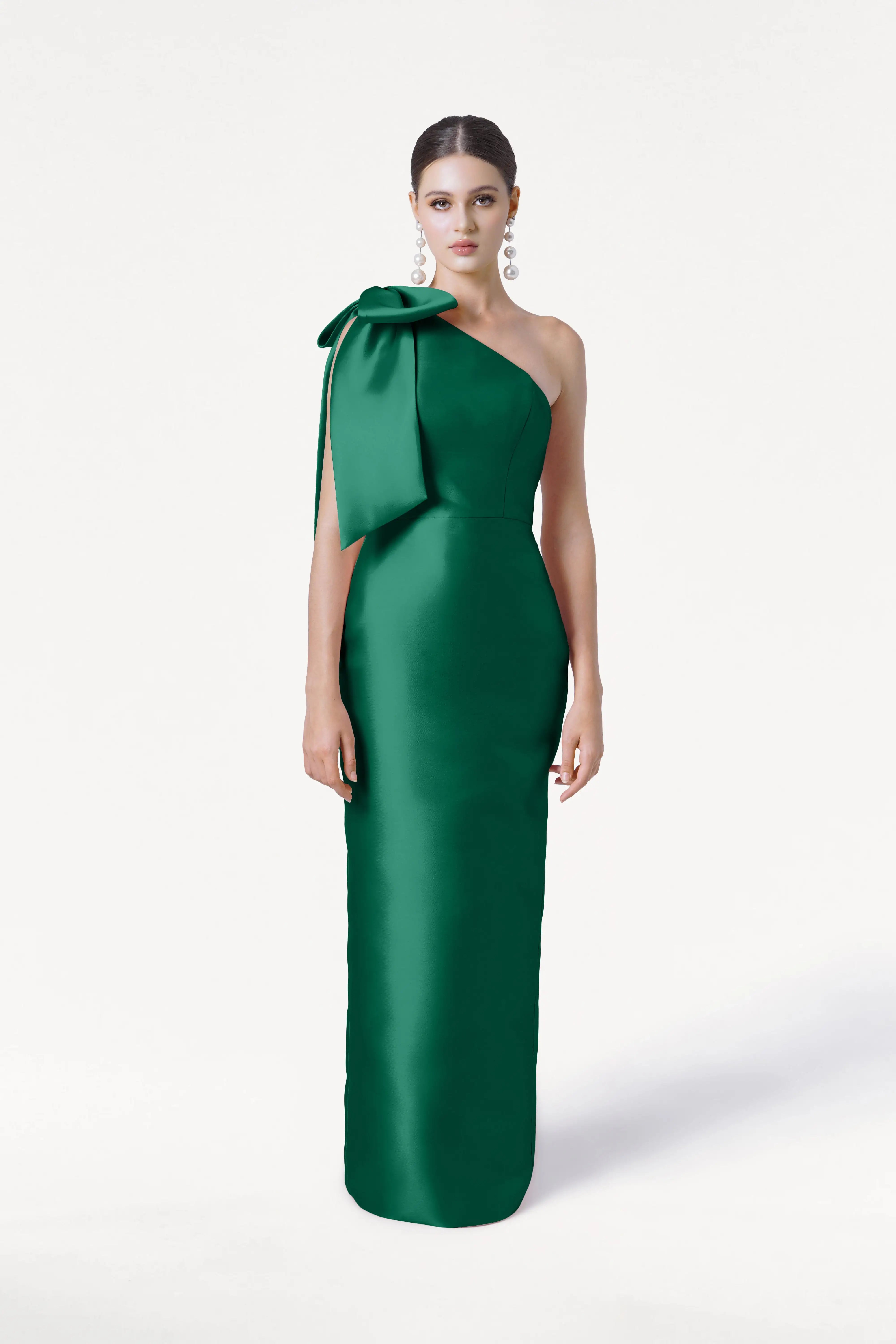 A woman stands poised against a white background wearing the TH&TH Ritz Bridesmaid Dress in Forest Green Satin Twill, a sleek, one-shoulder gown with a statement bow. Her hair is pulled back and she accessorizes with statement earrings.