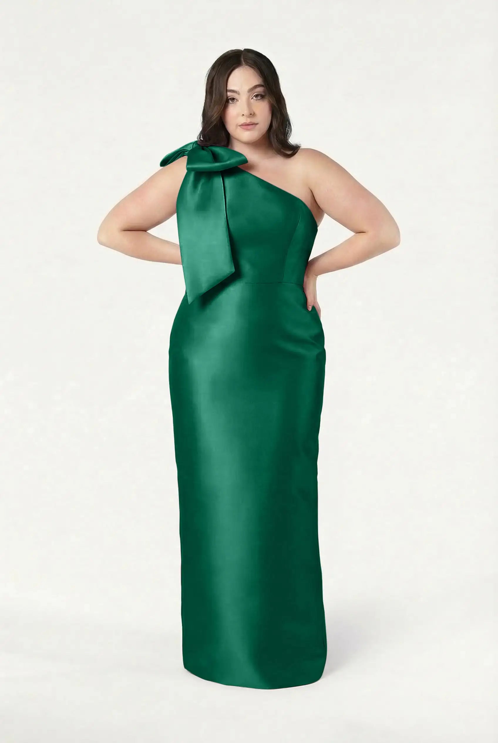 A woman poses with hands on hips in the TH&TH Ritz Bridesmaid Dress, a long, one-shoulder statement gown in forest green satin twill, against a plain white background.
