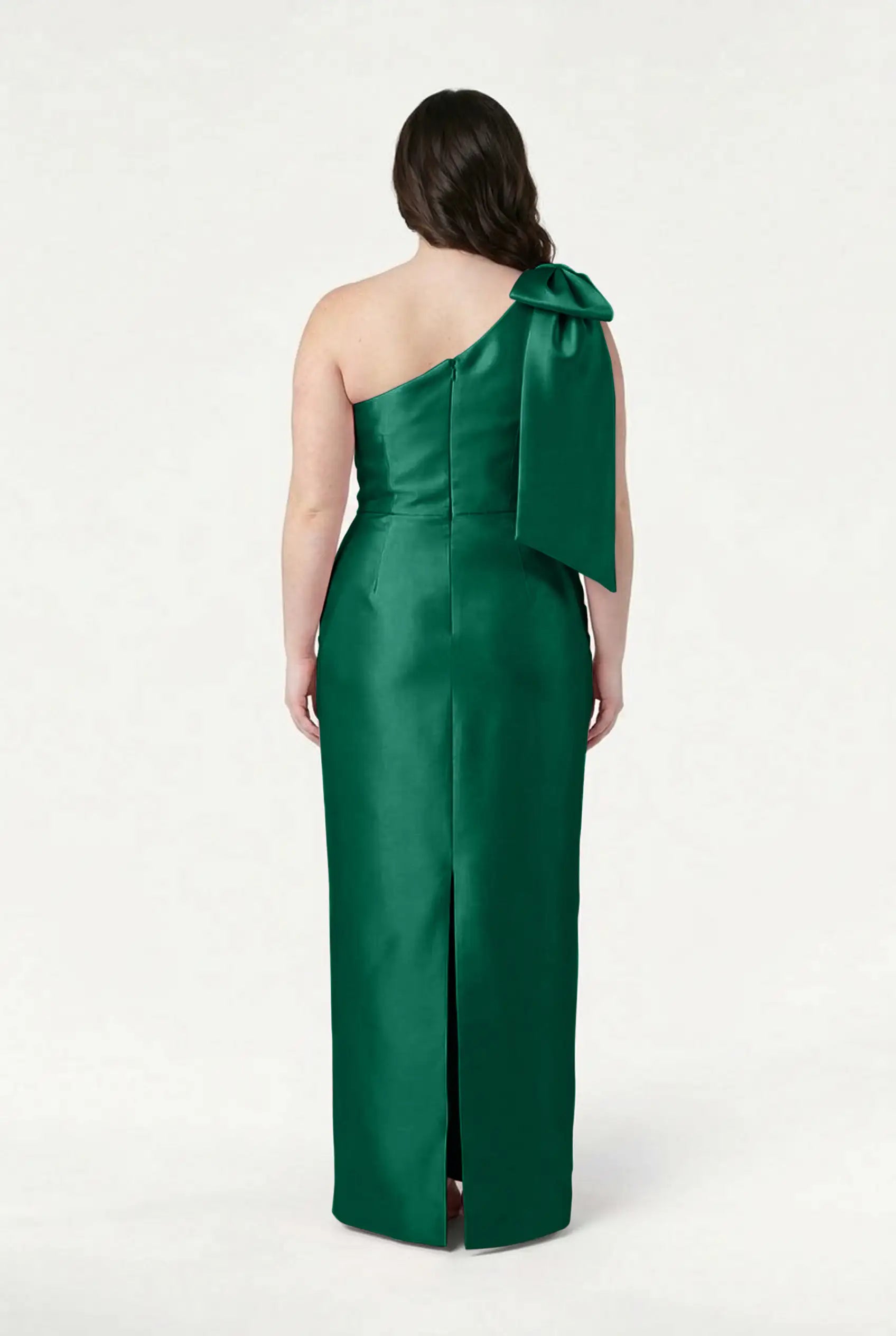 A woman in the TH&TH Ritz Bridesmaid Dress in Forest Green Satin Twill, featuring a one-shoulder design with a statement bow, stands facing away to showcase its elegant back and center hem slit.