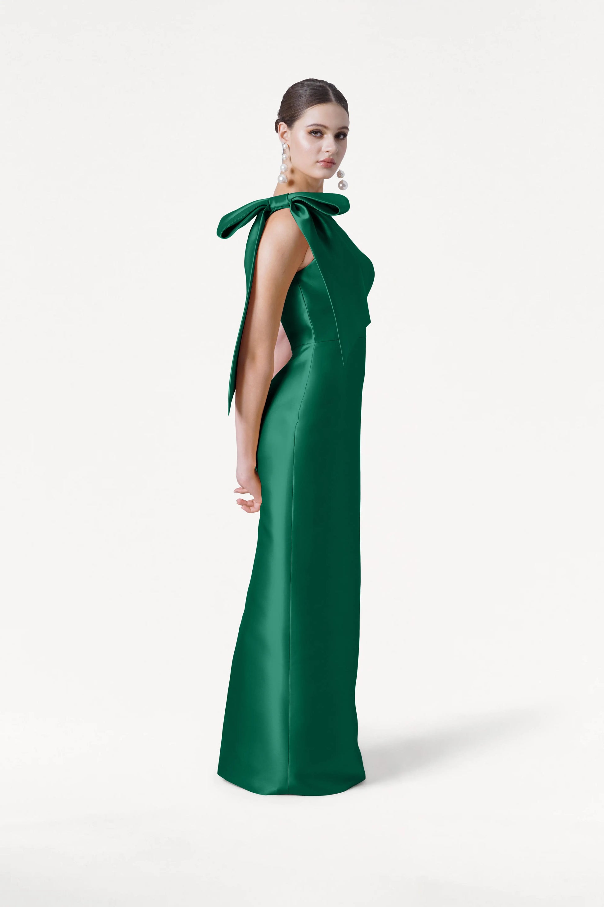 A woman in the TH&TH Ritz Bridesmaid Dress in Forest Green Satin Twill stands sideways against a plain white background. The floor-length, one-shoulder gown features a statement bow; her hair is pulled back and she wears statement earrings.