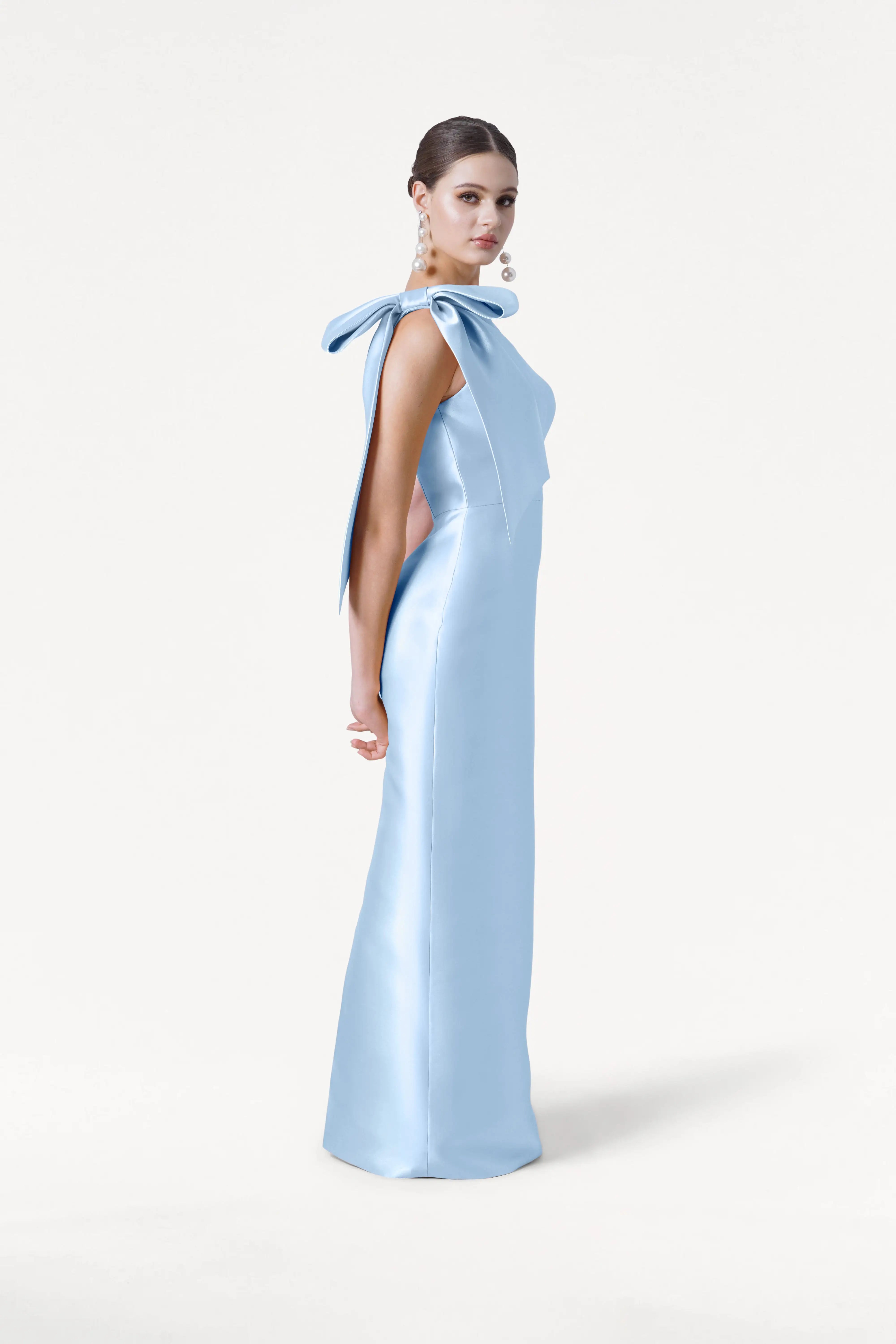 A woman stands in profile against a white backdrop wearing the TH&TH Ritz Bridesmaid Dress in Frosted Blue Satin Twill, featuring a floor-length silhouette, statement shoulder bow, open sides, slicked-back hair, and large spherical earrings.