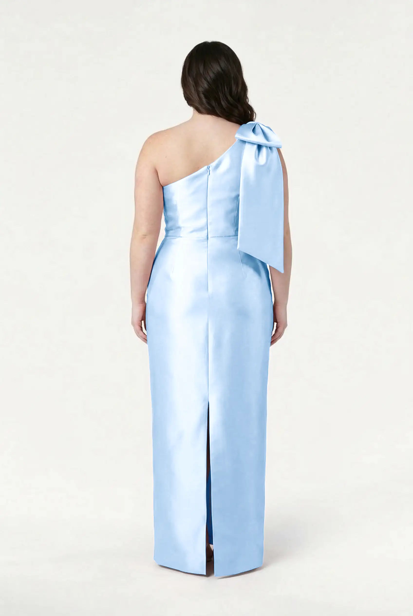 A woman with loose waves faces away, wearing the TH&TH Ritz Bridesmaid Dress in Frosted Blue Satin Twill. The one-shoulder style features a statement bow and back slit, set against a plain light background.