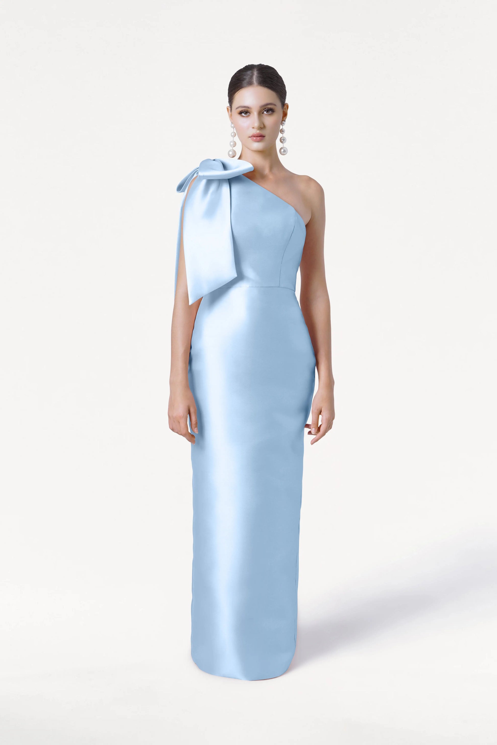A woman models the TH&TH Ritz Bridesmaid Dress in Frosted Blue Satin Twill, accented by a statement shoulder bow. Her hair is pulled back and she pairs the look with large dangling earrings against a plain white background.