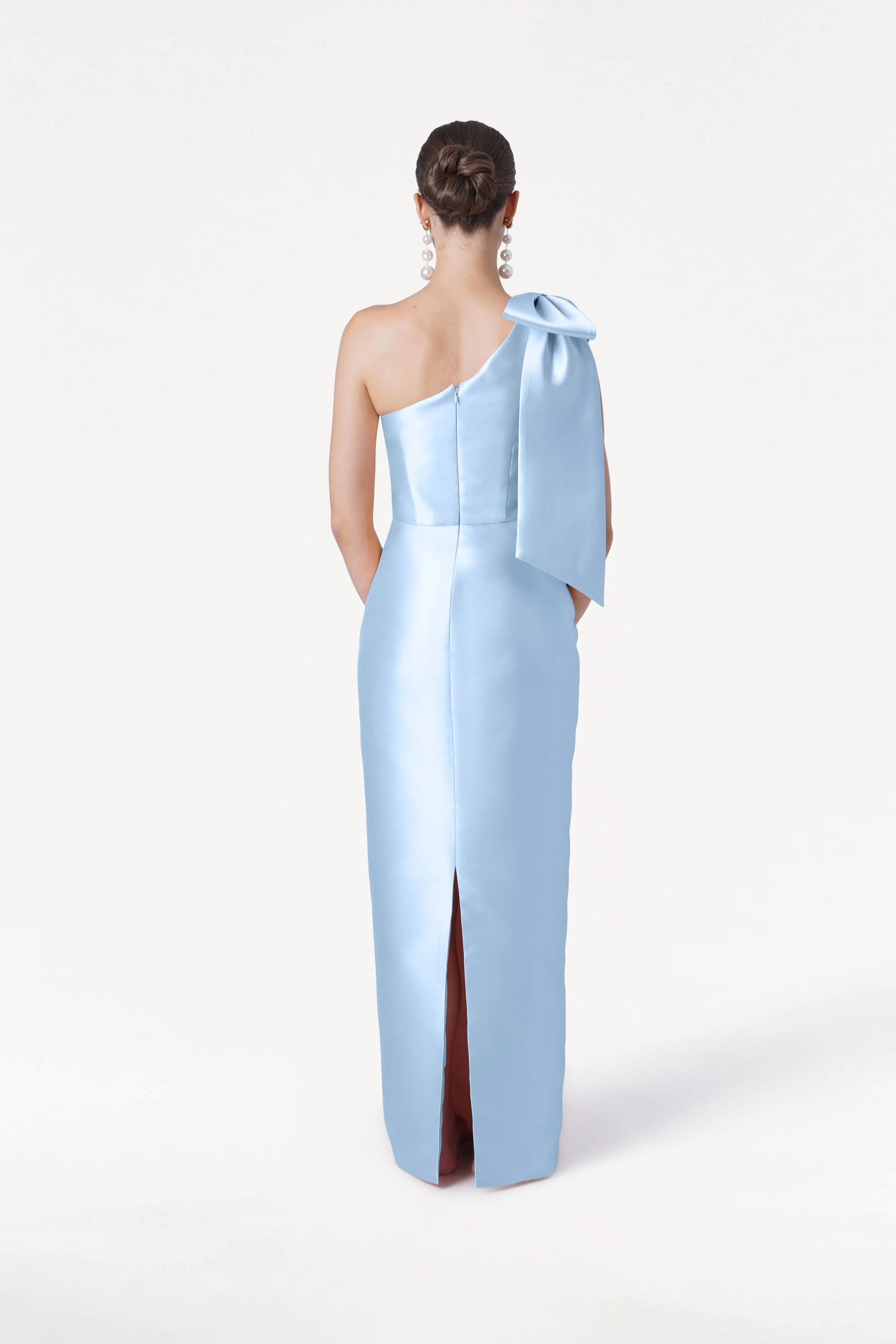 A woman faces away from the camera, wearing the TH&TH Ritz Bridesmaid Dress in Frosted Blue Satin Twill with a statement one-shoulder bow and back slit. Her hair is in a low bun with large earrings, set against a white background.
