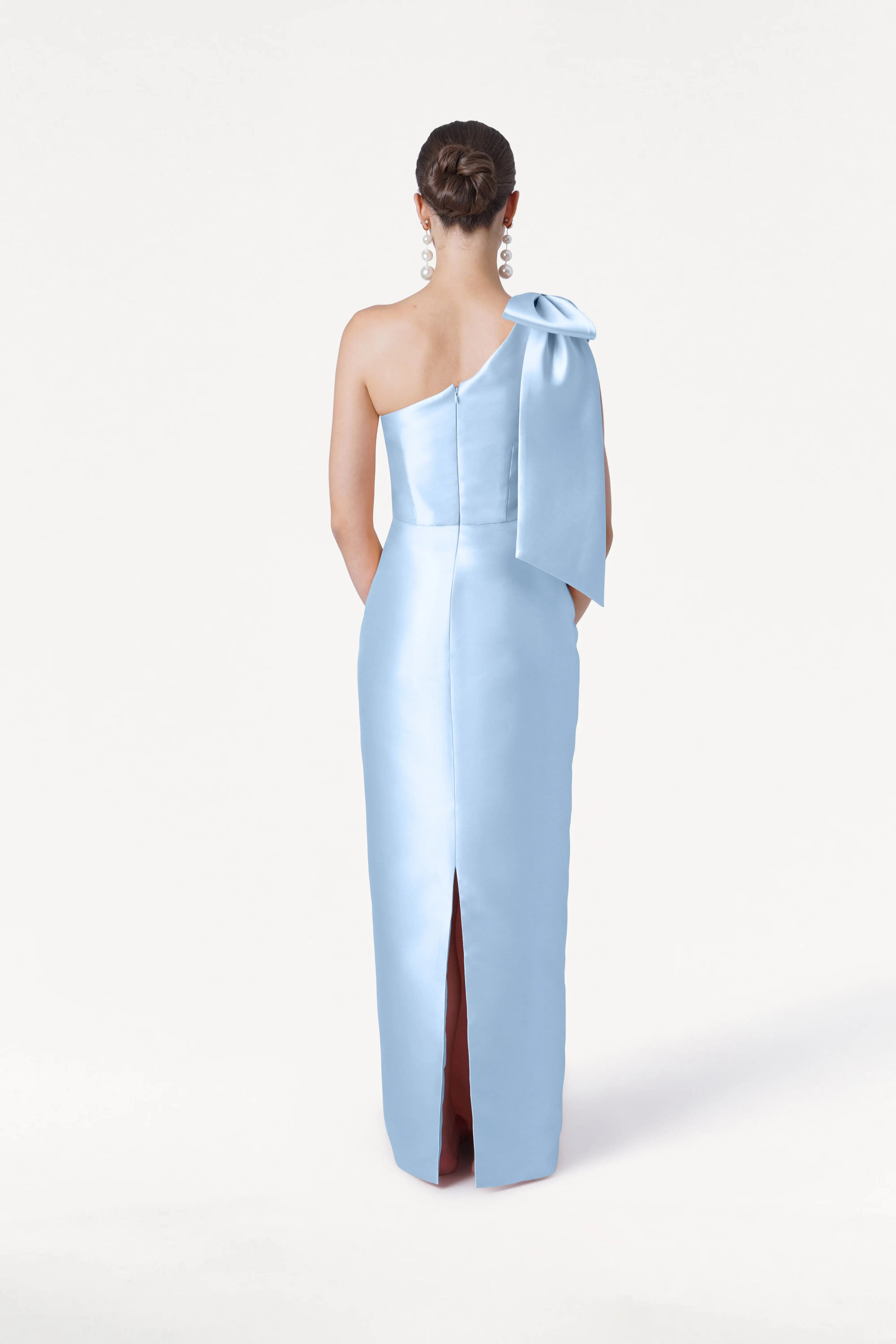 A woman faces away from the camera, wearing the TH&TH Ritz Bridesmaid Dress in Frosted Blue Satin Twill with a statement one-shoulder bow and back slit. Her hair is in a low bun with large earrings, set against a white background.