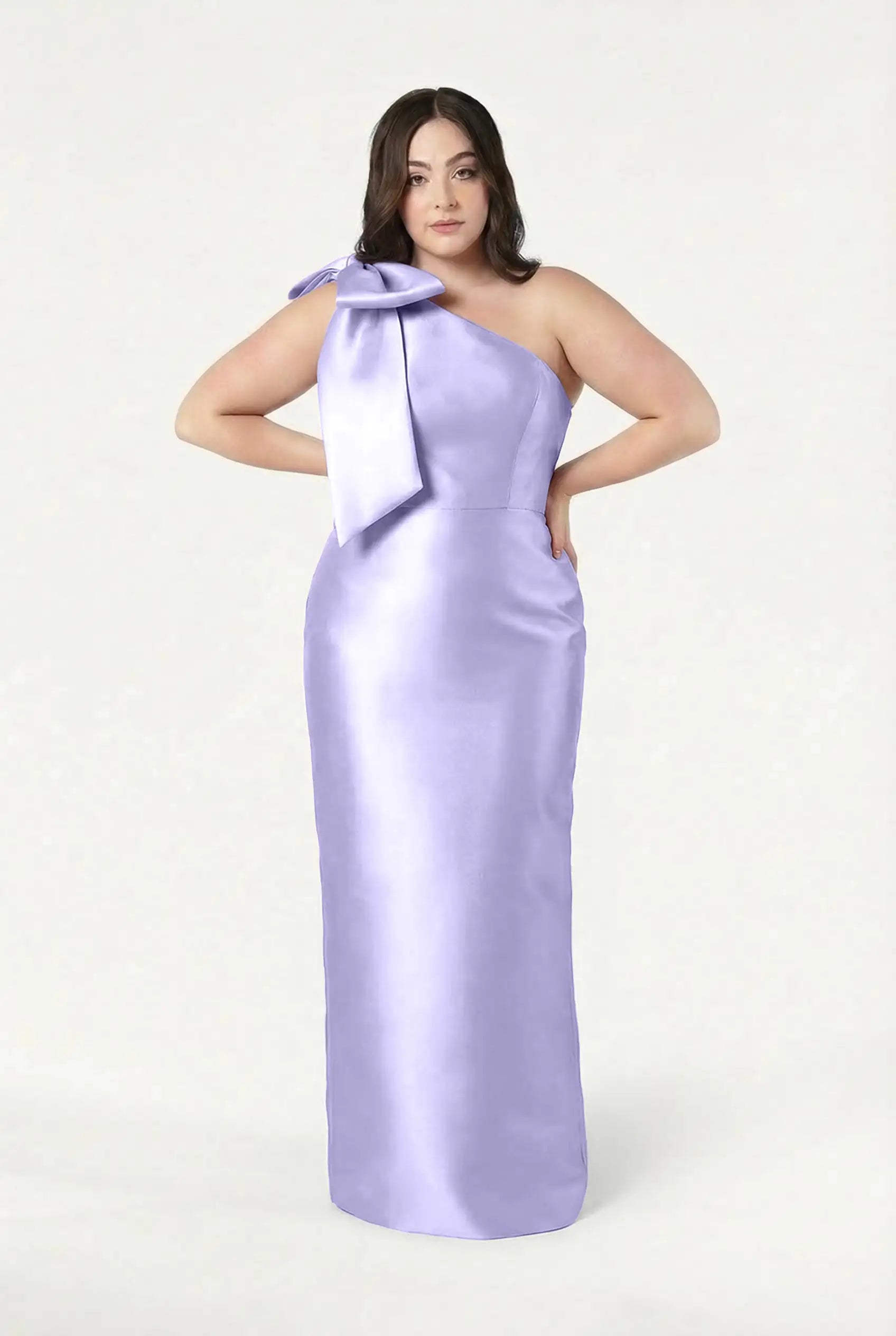 A woman poses with hands on hips in the TH&TH Ritz Bridesmaid Dress in Lilac Mist Satin Twill, featuring a one-shoulder design and statement bow, set against a plain light background.