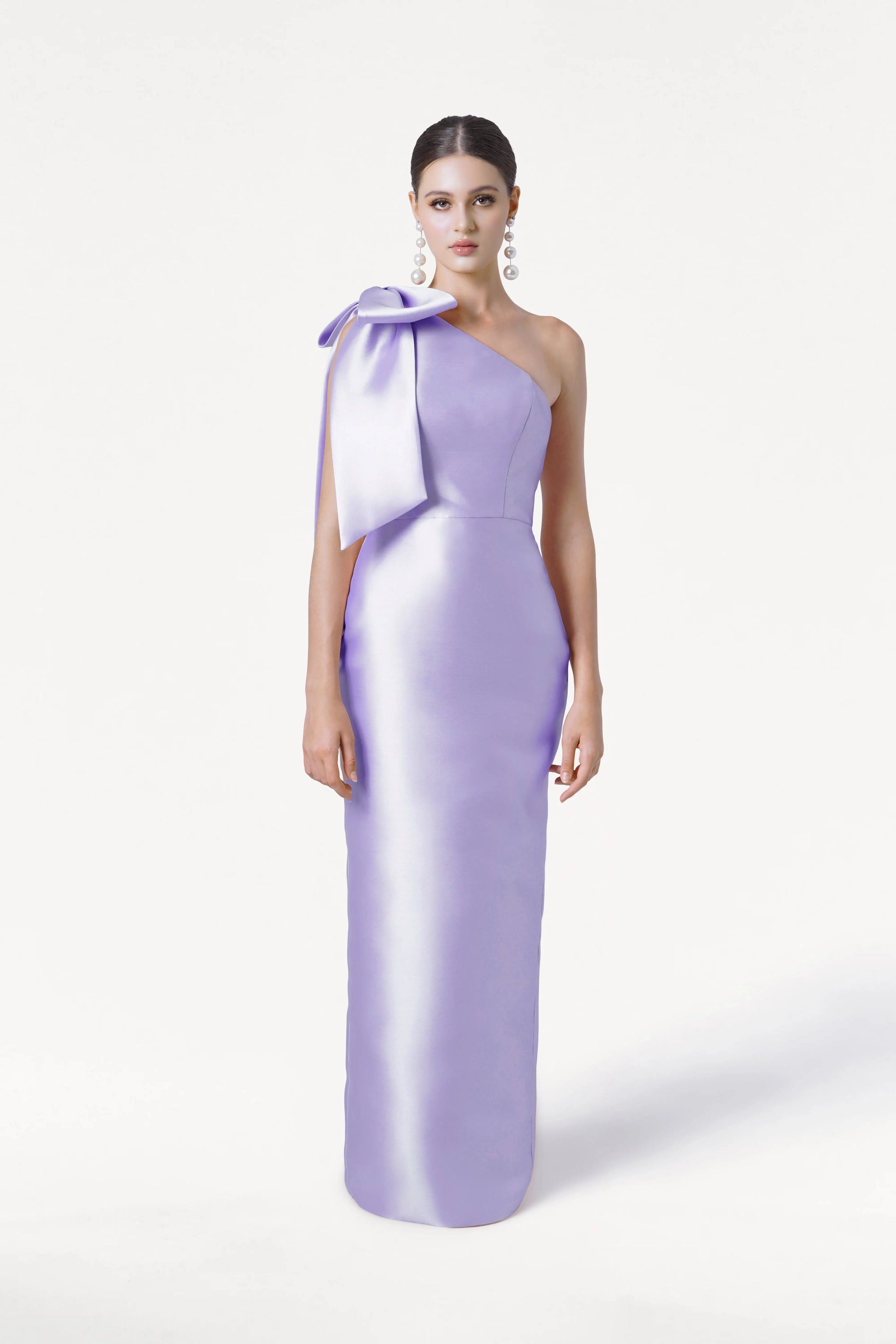 A woman models the TH&TH Ritz Bridesmaid Dress in Lilac Mist Satin Twill, featuring a one-shoulder design with a statement bow, against a white background. She wears bold earrings and has her hair slicked back.