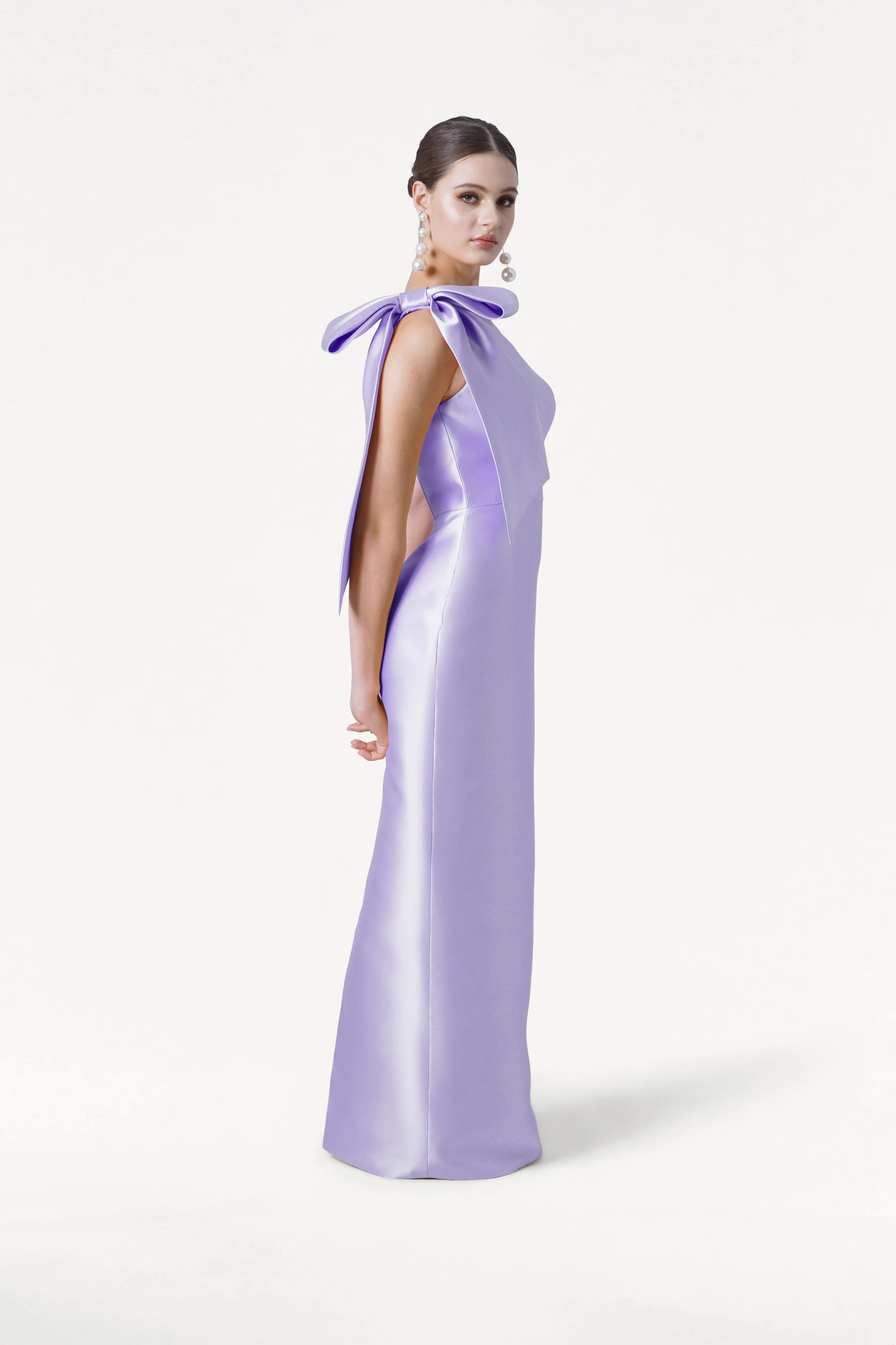 A woman stands sideways in the TH&TH Ritz Bridesmaid Dress in Lilac Mist Satin Twill, featuring a statement shoulder bow. Her hair is styled back, and she wears large pearl earrings against a plain white background.