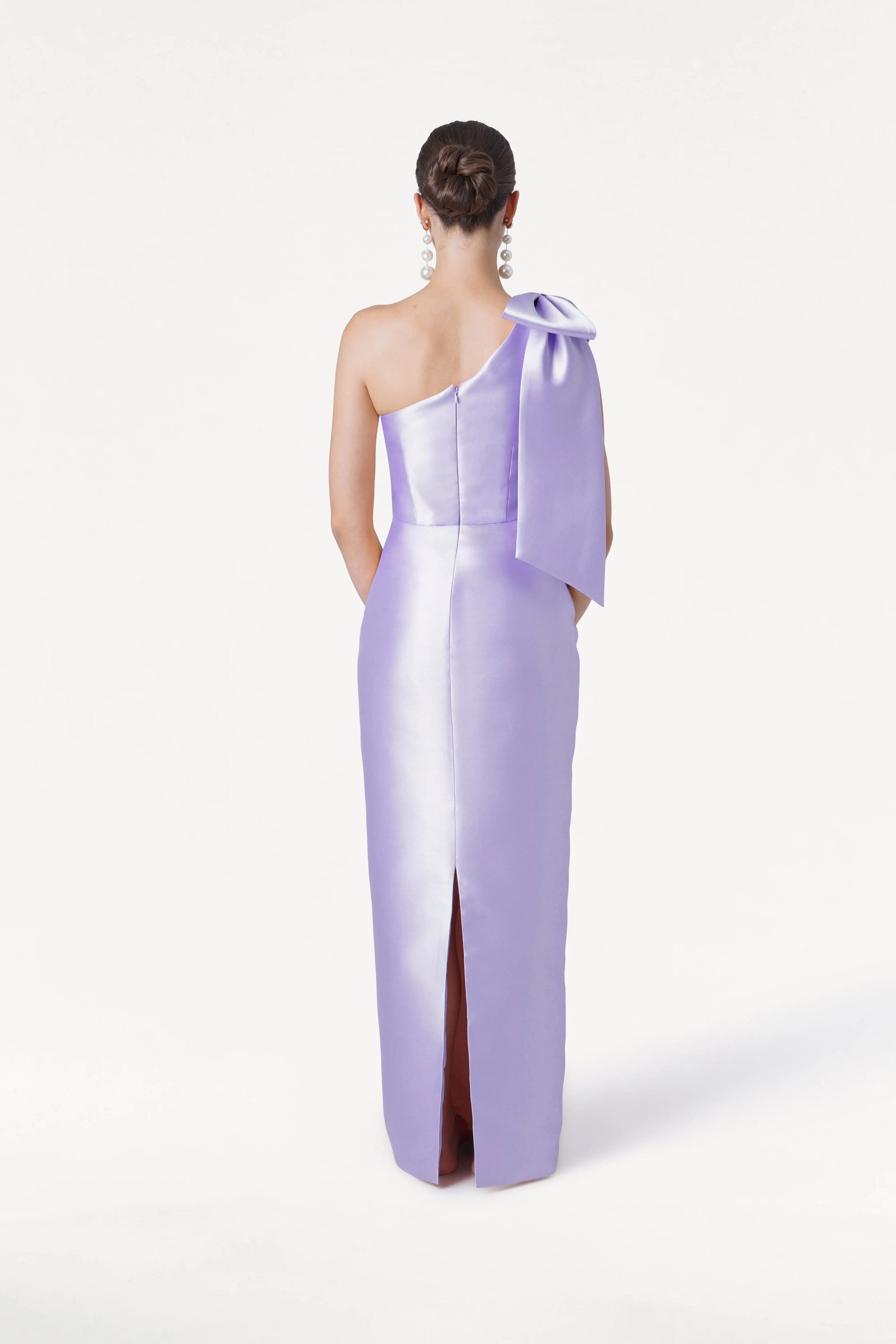 A woman with her hair in a neat bun and dangling earrings models the TH&TH Ritz Bridesmaid Dress in Lilac Mist Satin Twill, featuring a one-shoulder design, statement shoulder bow, and back slit, against a plain white background.