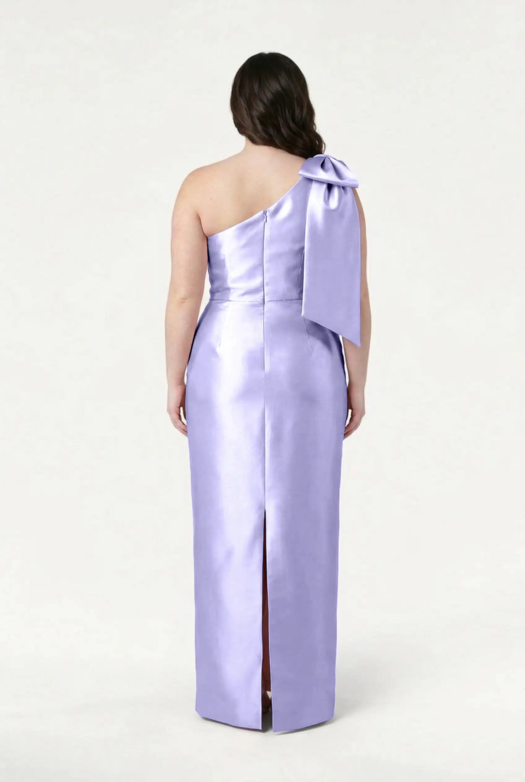 A woman faces away, showcasing the fitted silhouette and back slit of the TH&TH Ritz Bridesmaid Dress in Lilac Mist Satin Twill—a shiny, one-shoulder gown—against a plain light background.