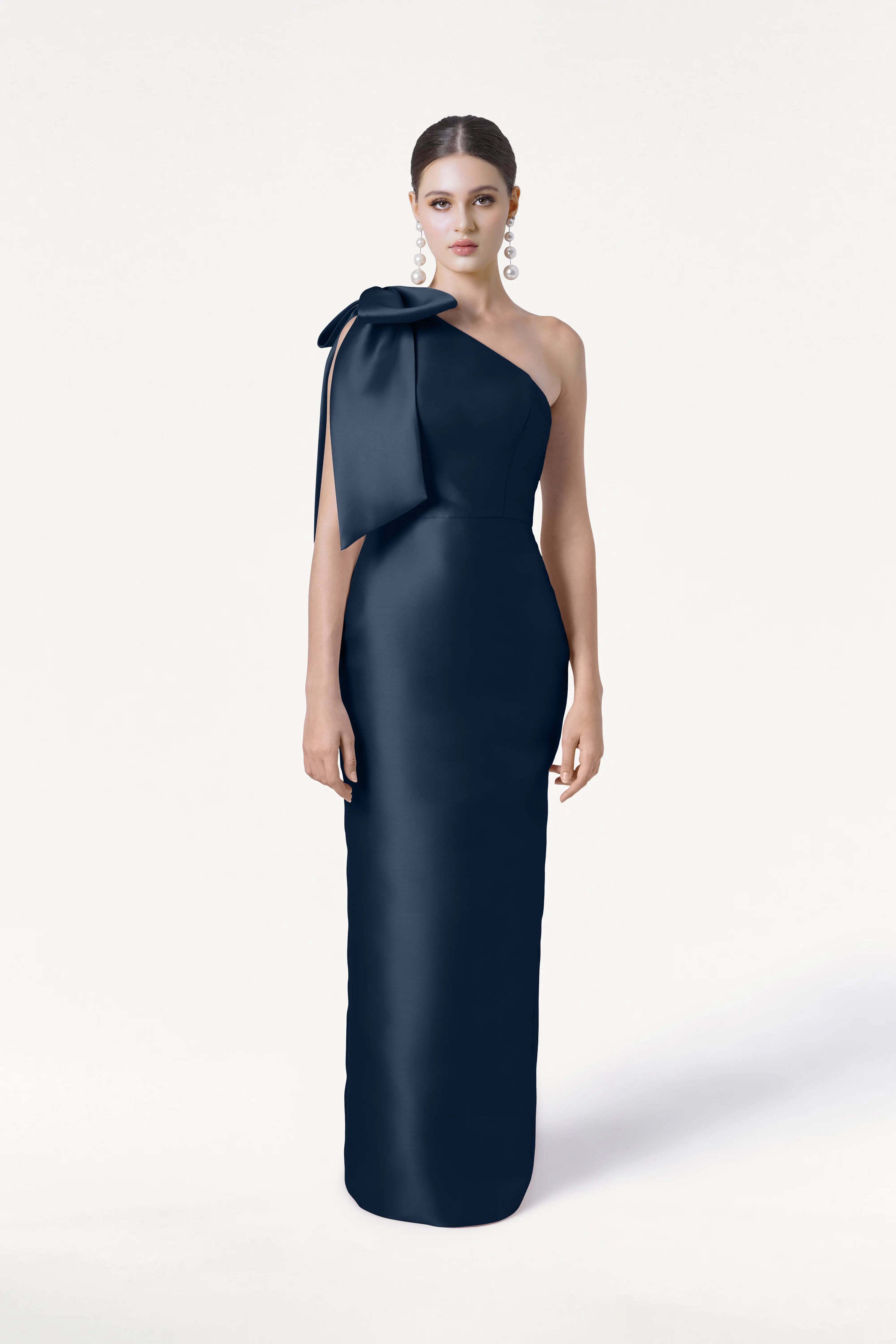 A woman wears the TH&TH Ritz Bridesmaid Dress in Navy Satin Twill, featuring a one-shoulder design with a statement bow. Her hair is pulled back and she pairs the look with long earrings against a plain white background.