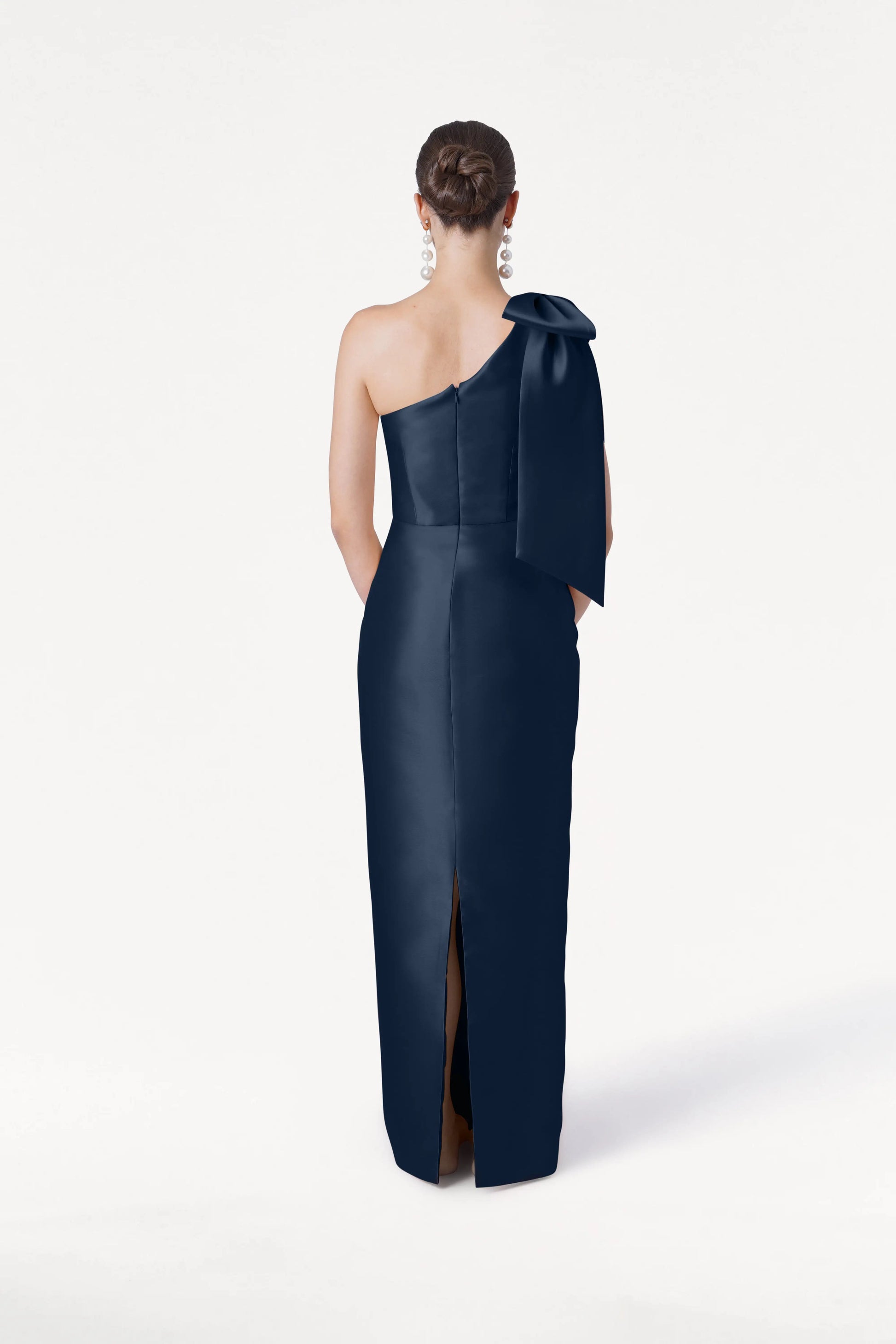 A woman stands with her back to the camera, wearing the TH&TH Ritz Bridesmaid Dress in Navy Satin Twill. The gown features a one-shoulder silhouette, statement bow, and back slit. She accessorizes with dangling earrings against a white background.