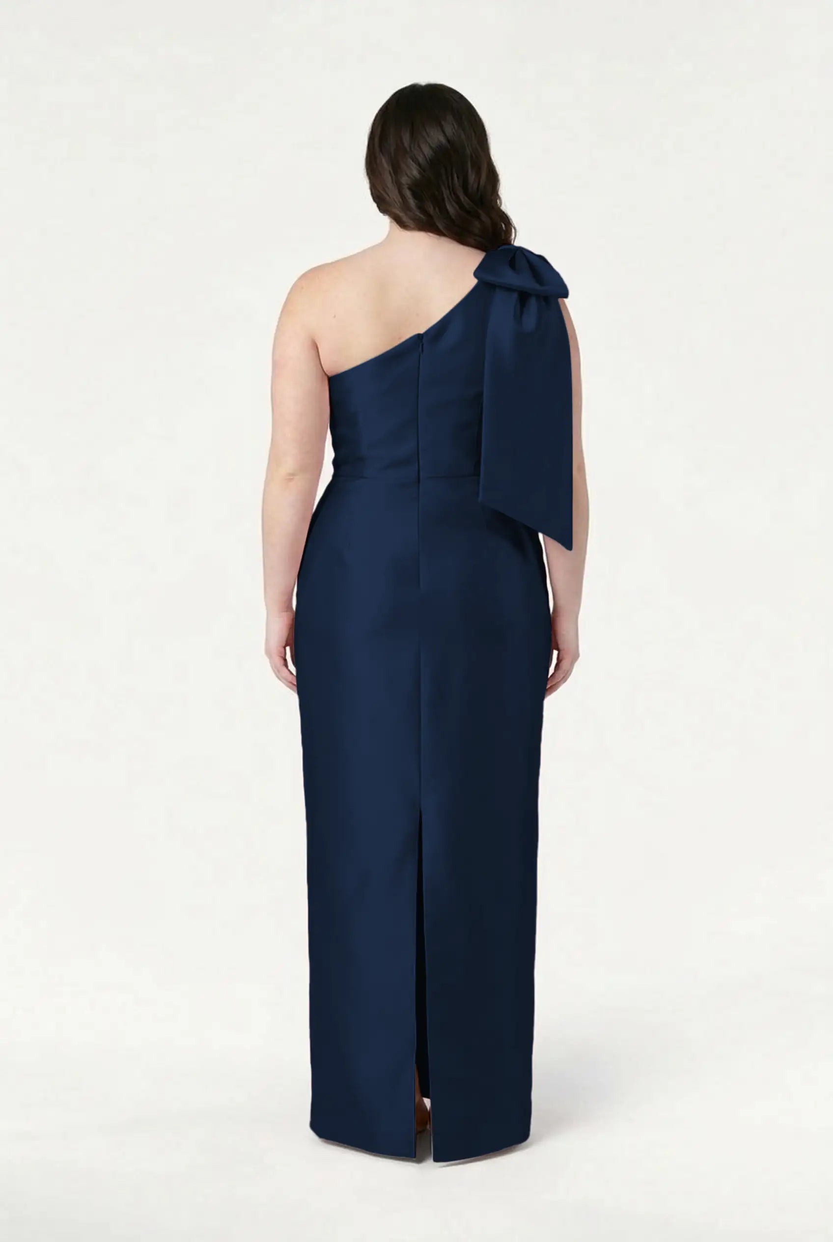 A woman models the TH&TH Ritz Bridesmaid Dress in Navy Satin Twill, standing with her back to the camera to highlight the one-shoulder design, statement bow, straight skirt, and back slit against a plain white background.