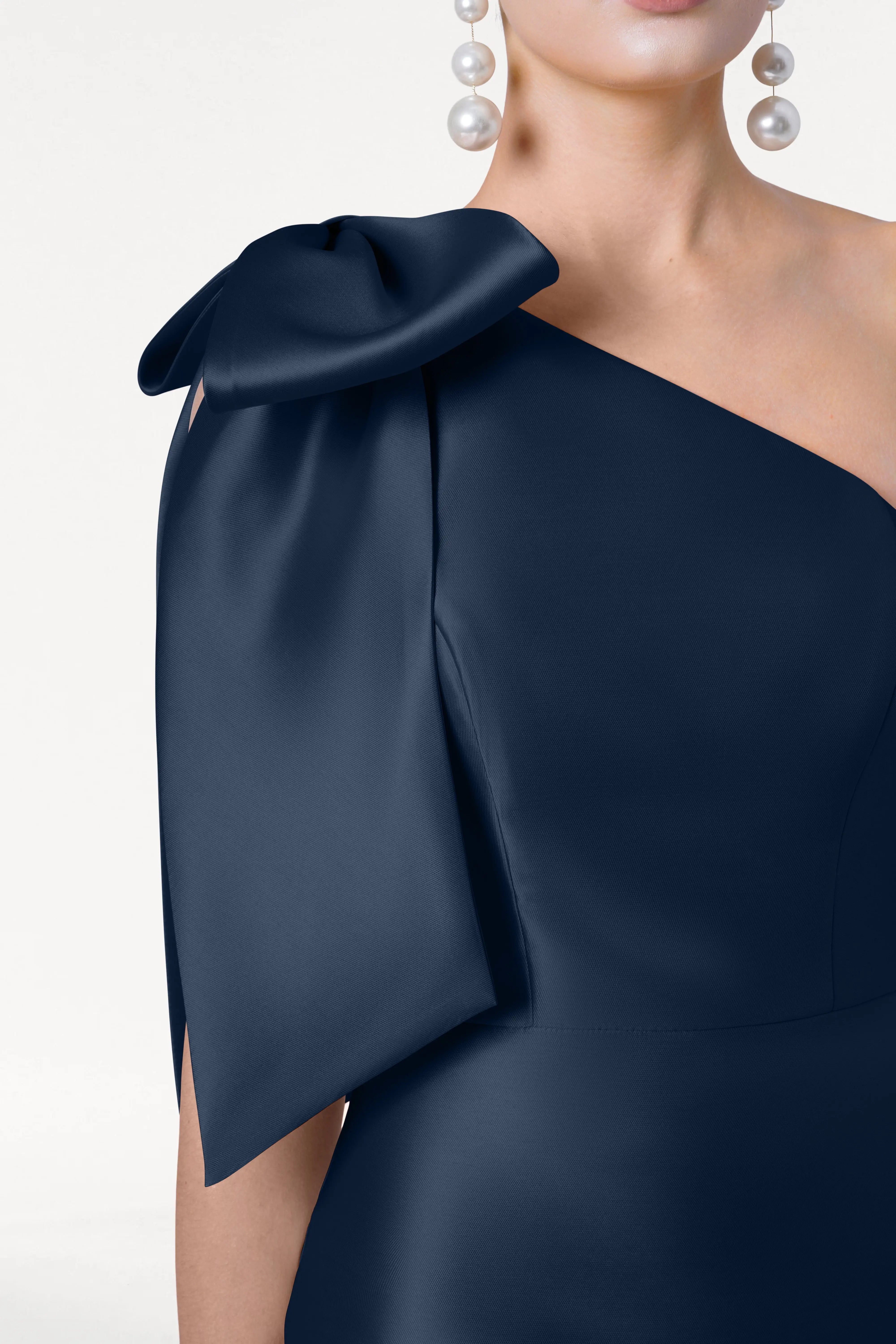 A woman models the TH&TH Ritz Bridesmaid Dress in Navy Satin Twill, featuring a one-shoulder design with a statement bow, paired with drop pearl earrings, shown from neck to waist against a plain white background.