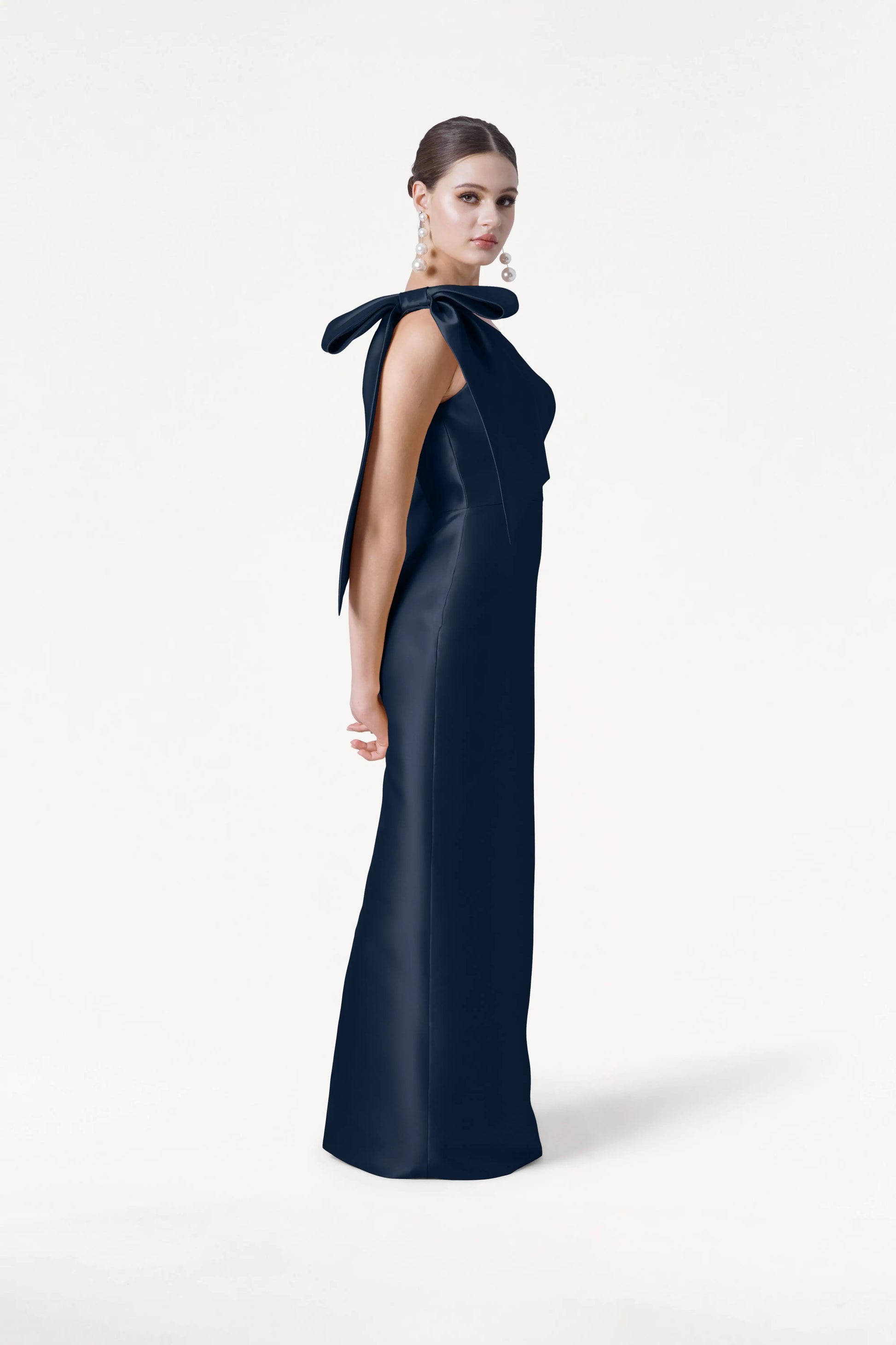 A woman in profile wears the TH&TH Ritz Bridesmaid Dress in Navy Satin Twill, featuring a statement bow on one shoulder. Her hair is styled back, complemented by statement earrings, against a plain white background.