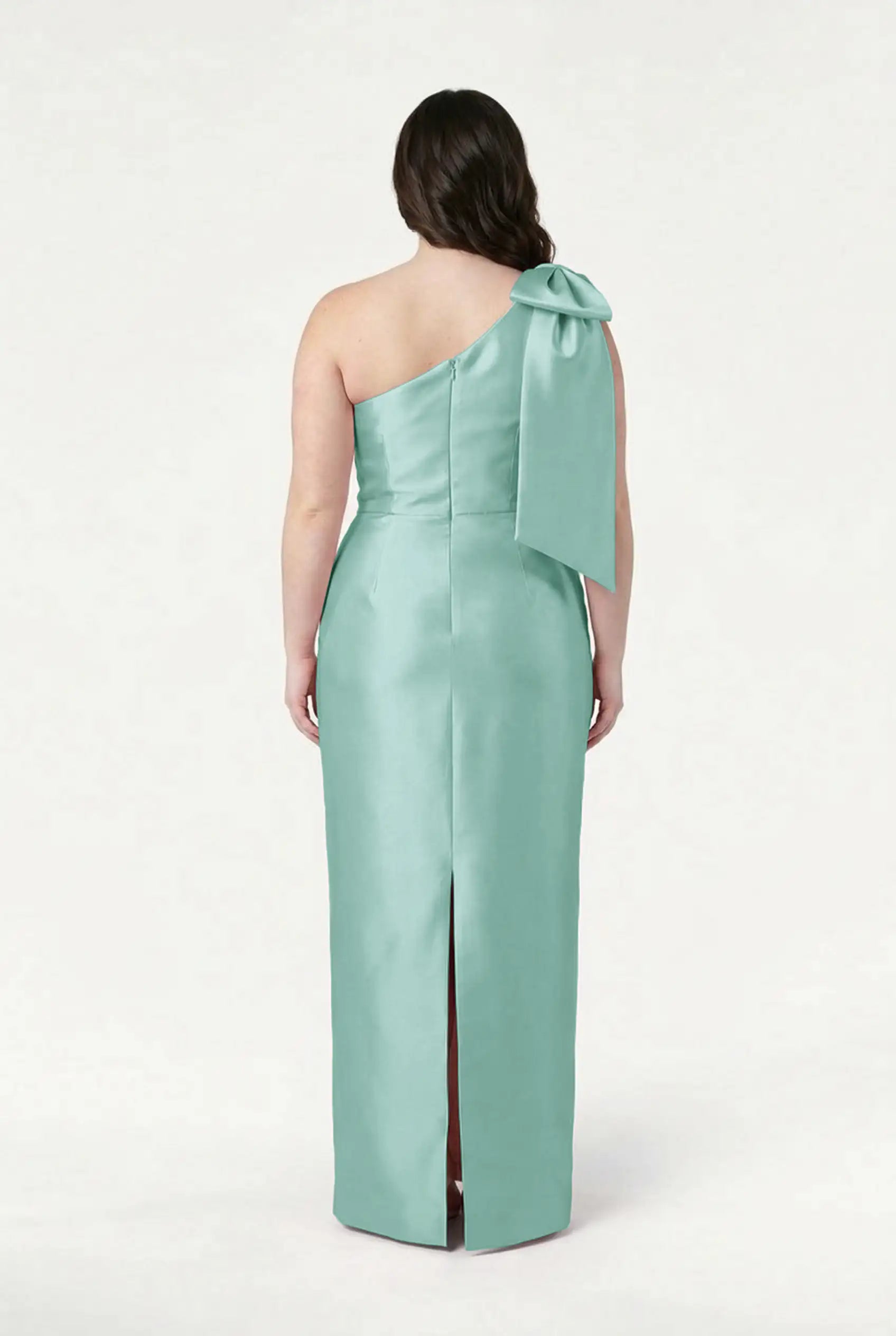 A person faces away, wearing the TH&TH Ritz Bridesmaid Dress in Seafoam Satin Twill—a one-shoulder gown with a large statement shoulder bow and back slit—against a plain white background.