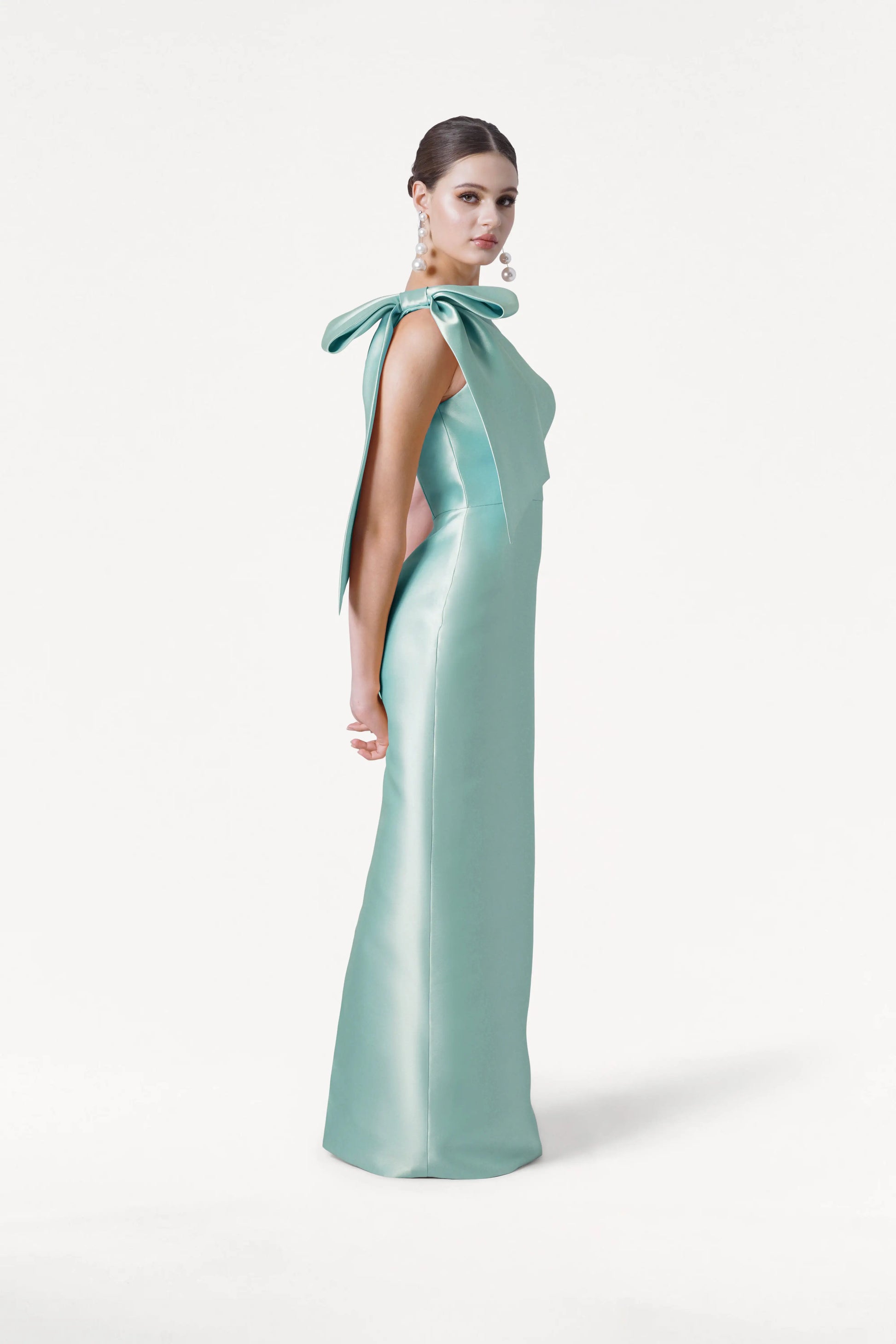 A woman wears the TH&TH Ritz Bridesmaid Dress in Seafoam Satin Twill, featuring a floor-length silhouette and a statement one-shoulder bow. She stands against a white background, hair styled back, accessorized with large dangling earrings.