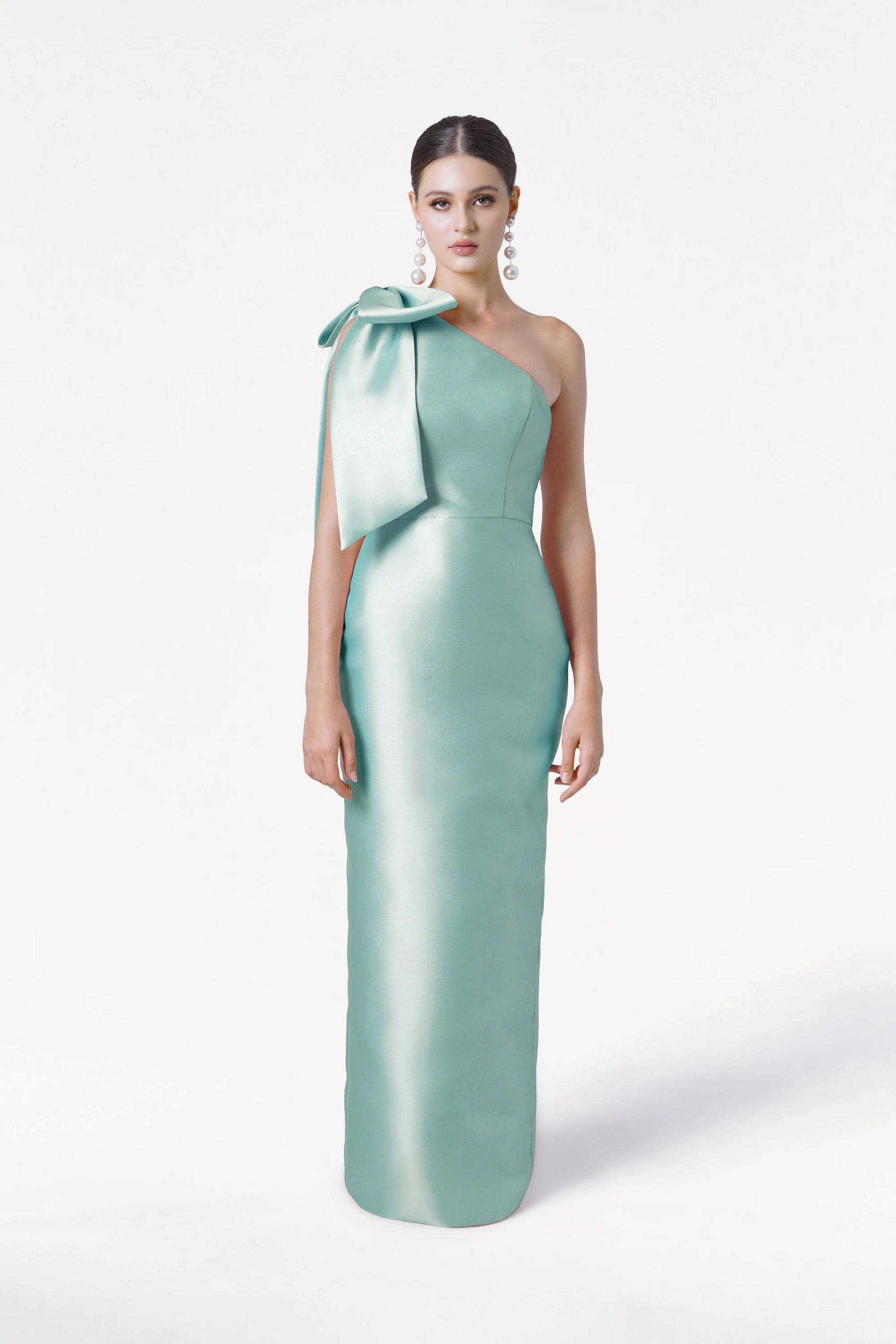 A woman models the TH&TH Ritz Bridesmaid Dress in Seafoam Satin Twill, a shiny one-shoulder gown with a statement bow. Her hair is slicked back and she wears large earrings, set against a plain white background.