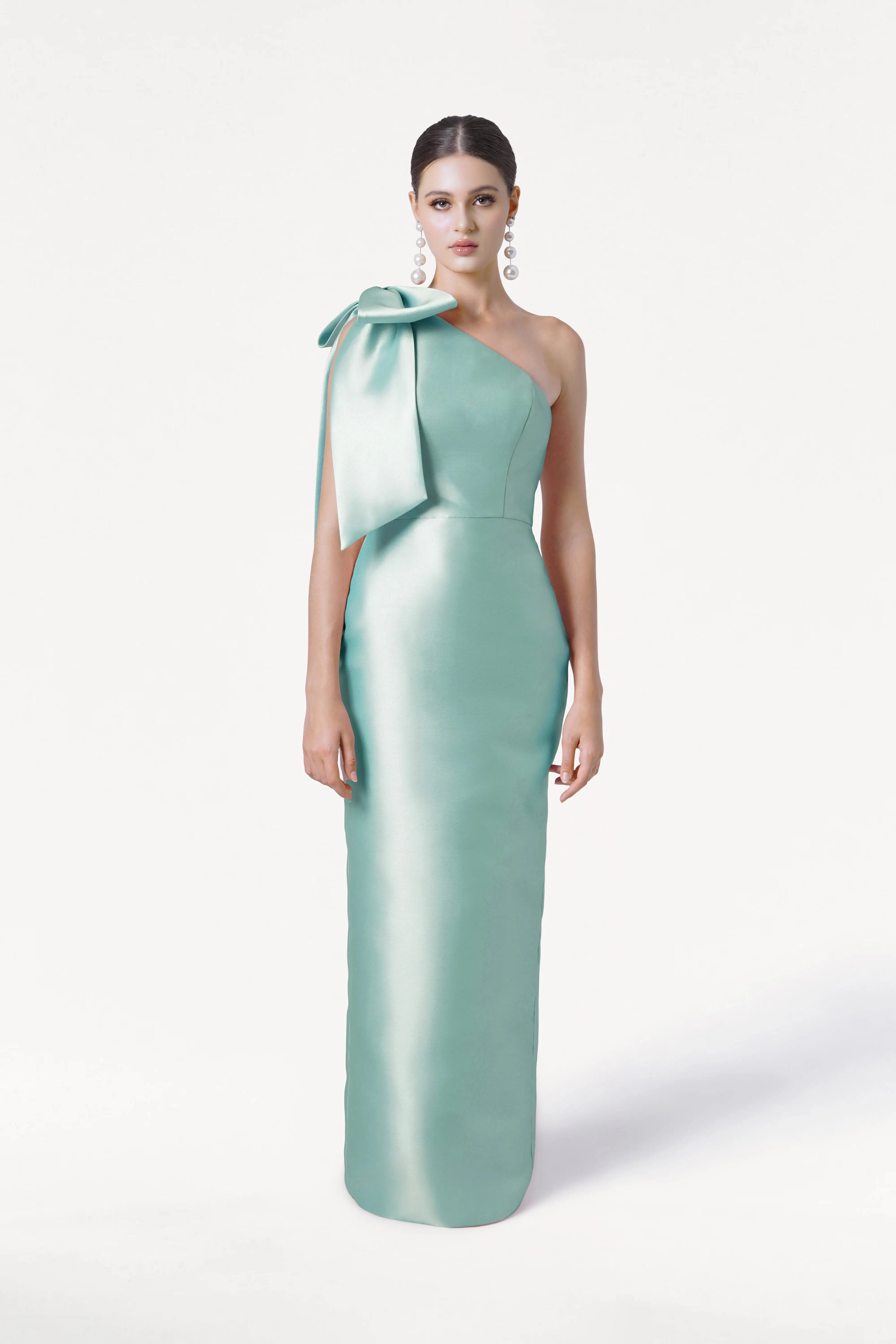 A woman models the TH&TH Ritz Bridesmaid Dress in Seafoam Satin Twill, a shiny one-shoulder gown with a statement bow. Her hair is slicked back and she wears large earrings, set against a plain white background.