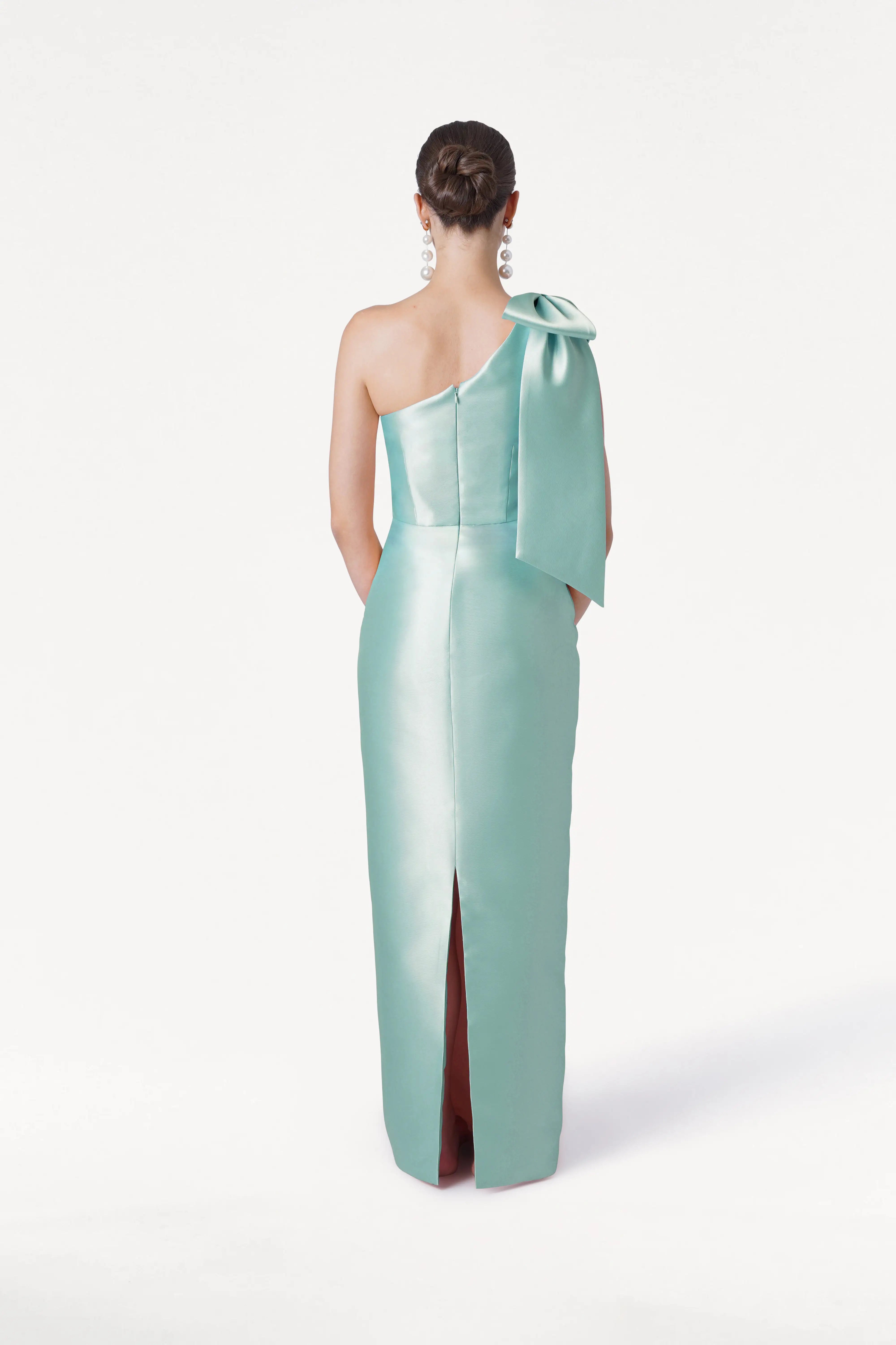 A woman with dark hair in a bun models the TH&TH Ritz Bridesmaid Dress in Seafoam Satin Twill—a shiny one-shoulder gown featuring a statement shoulder bow and back slit—against a plain white background.