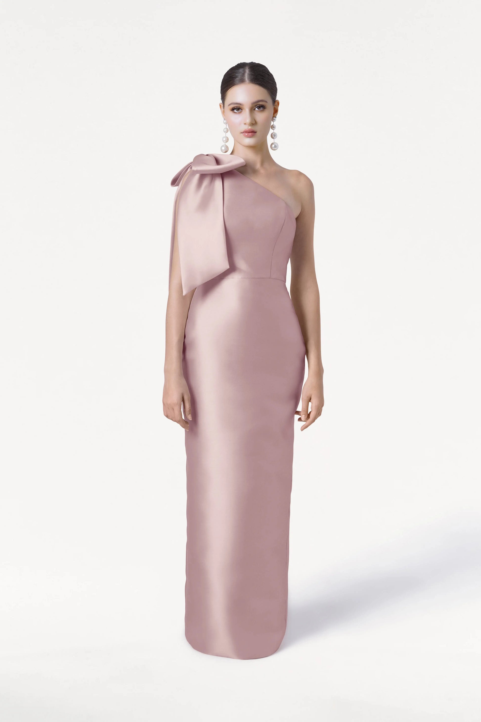 A woman with dark hair pulled back and large dangling earrings models the TH&TH Ritz Bridesmaid Dress in Smoked Blush Satin Twill, a long one-shoulder gown with a statement bow, against a plain white background.