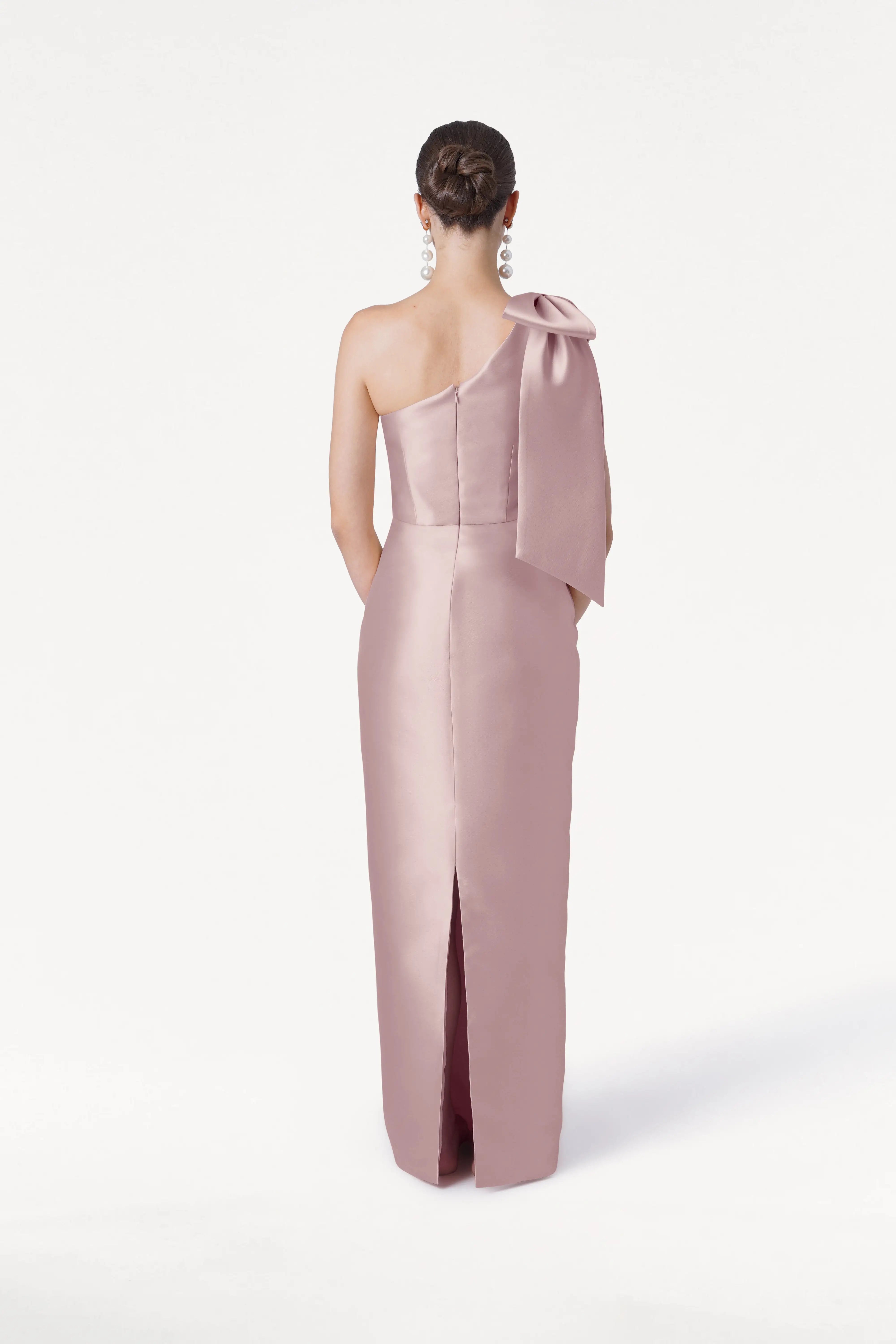 A woman faces away, wearing the TH&TH Ritz Bridesmaid Dress in Smoked Blush Satin Twill—a light pink one-shoulder gown with a statement bow and back slit. Her hair is in a low bun, with large earrings, set against a plain white background.