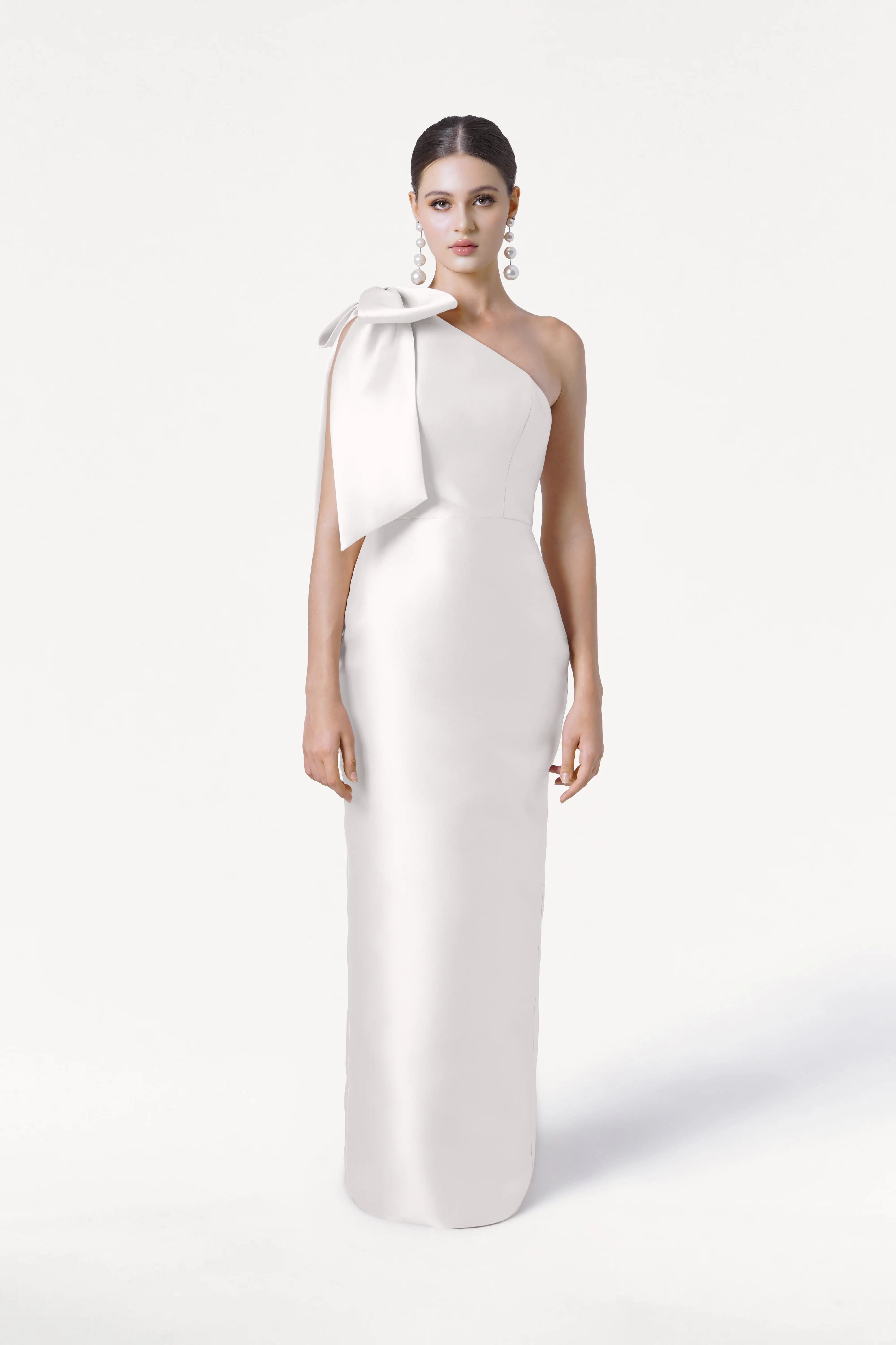 A woman models the TH&TH Ritz Dress in Ivory Satin Twill, a sleek, floor-length one-shoulder bridesmaid gown with a statement shoulder bow. She completes the modern bridal look with statement earrings and slicked-back hair against a white backdrop.
