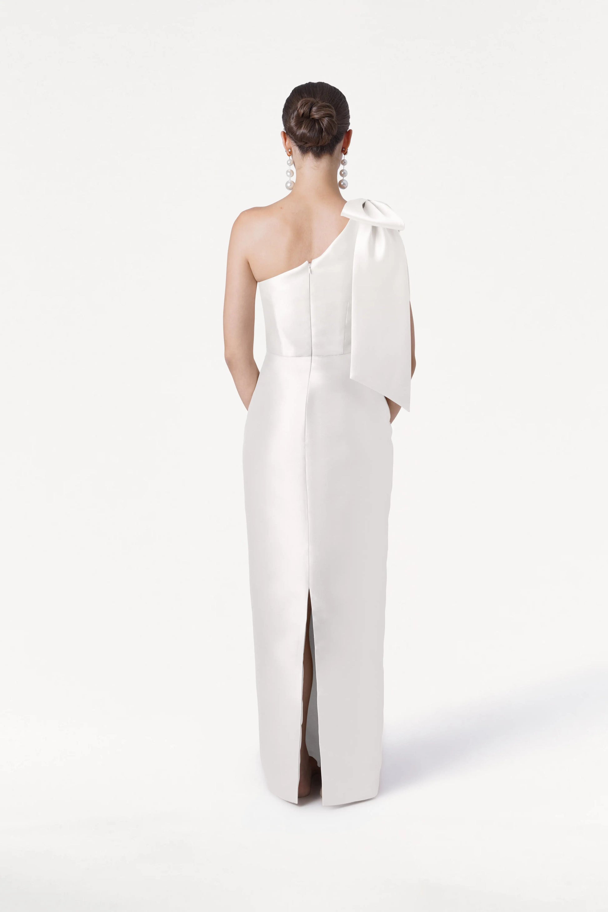 A woman faces away, wearing the TH&TH Ritz Dress in Ivory Satin Twill—a one-shoulder bridesmaid gown with a statement bow, back slit, and her hair in a neat bun. The plain white background enhances the modern bridal vibe.