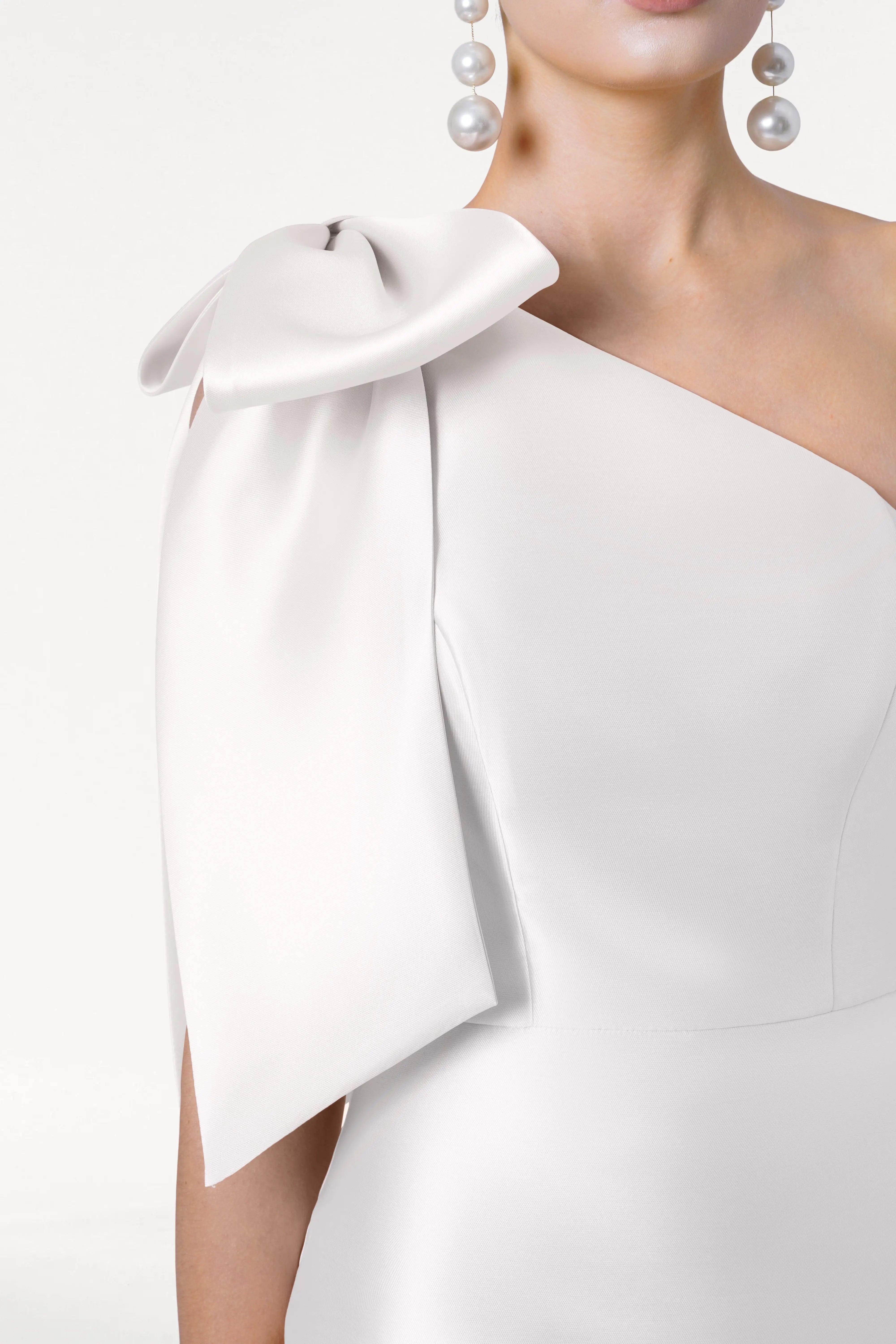 A woman models the TH&TH Ritz Dress in Ivory Satin Twill, featuring a one-shoulder design with a large bow and pearl earrings, shown from the waist up against a white background—ideal for modern bridal parties.