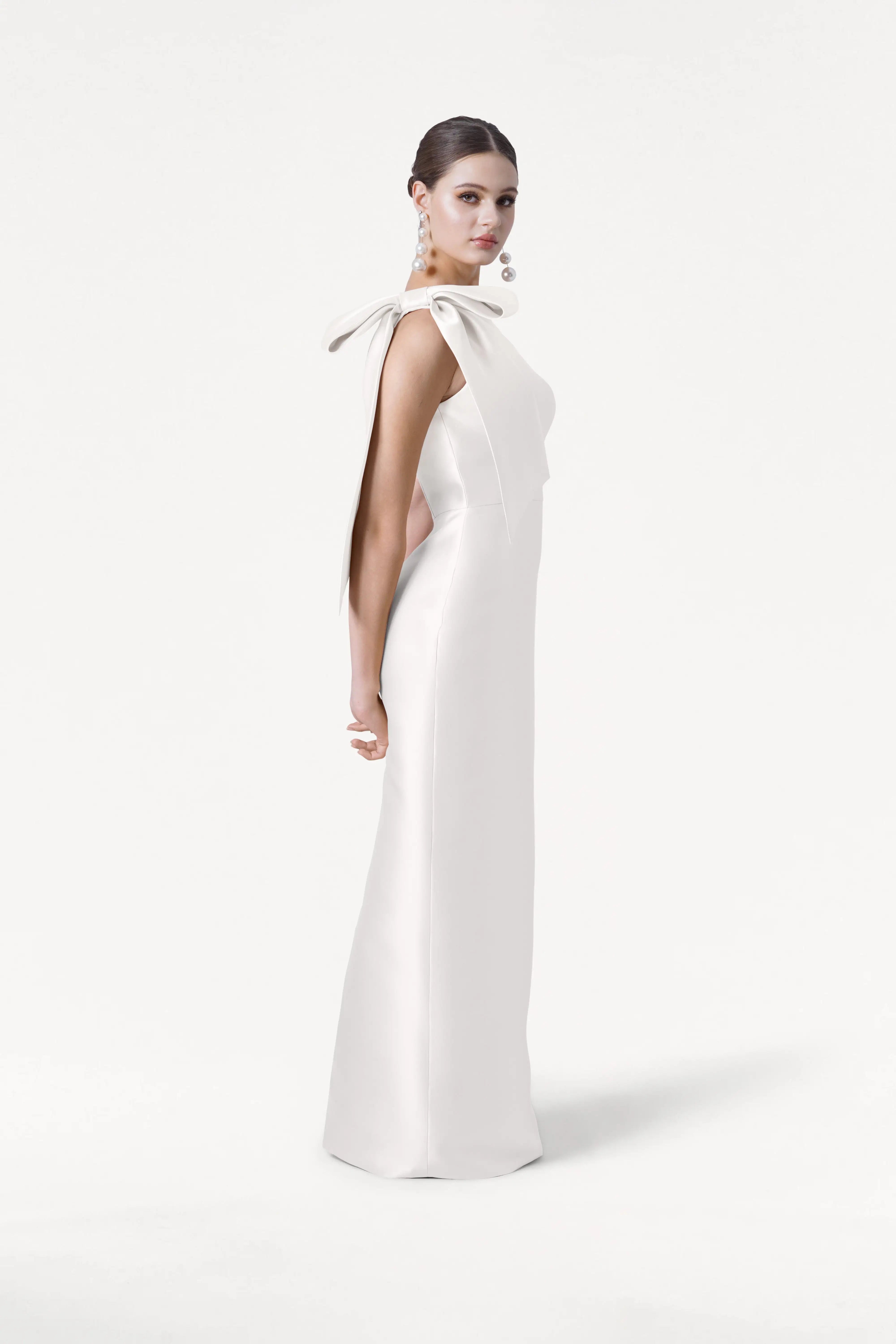 A woman in profile wears the TH&TH Ritz Dress in Ivory Satin Twill, featuring a one-shoulder design with a large bow. Her hair is styled back with statement earrings—a chic look for modern bridal parties against a white background.