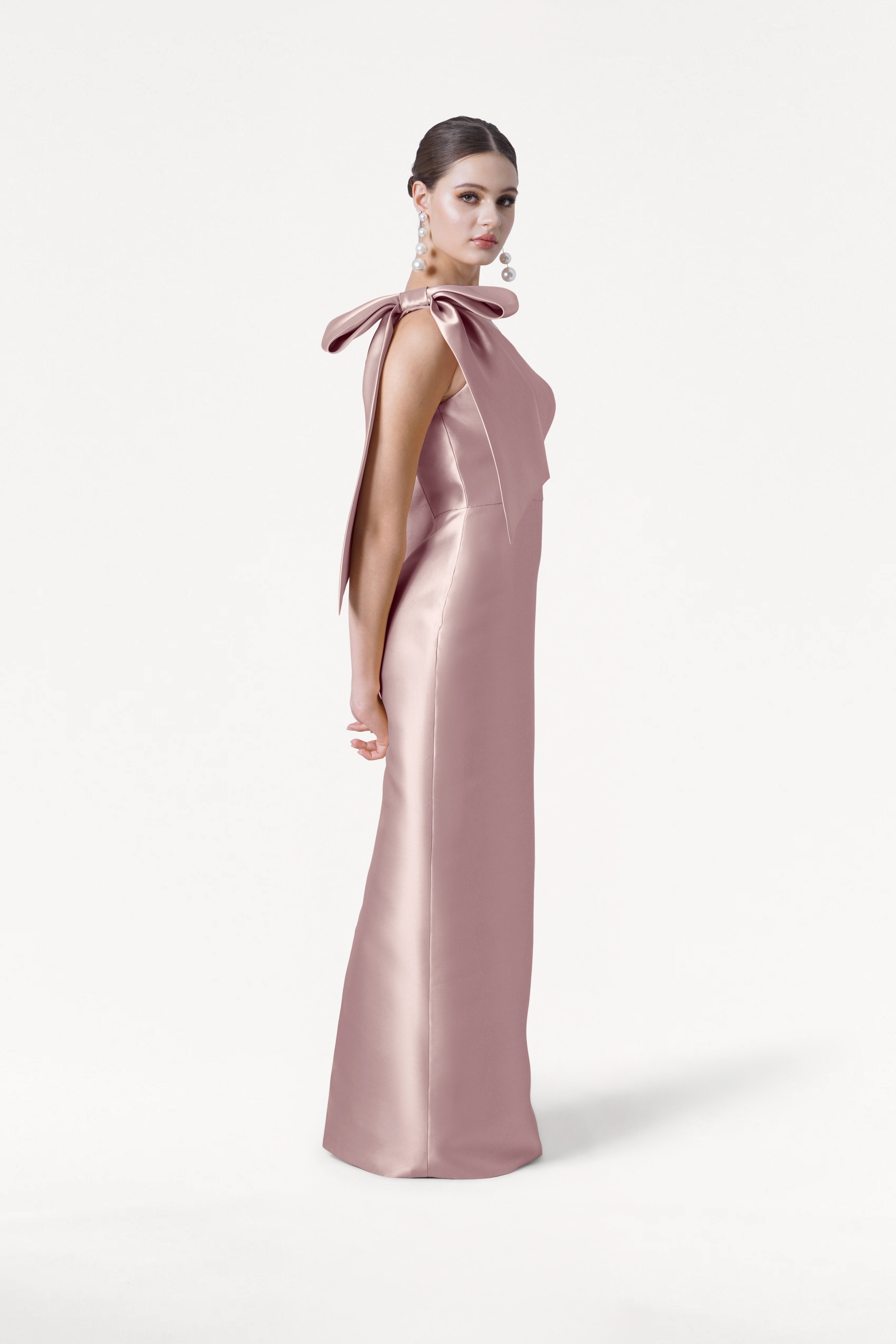 A woman poses in profile against a white background, wearing TH&TH's Ritz Bridesmaid Dress in Smoked Blush Satin Twill with a statement bow on one shoulder. Her hair is slicked back and she pairs bold earrings with the look.