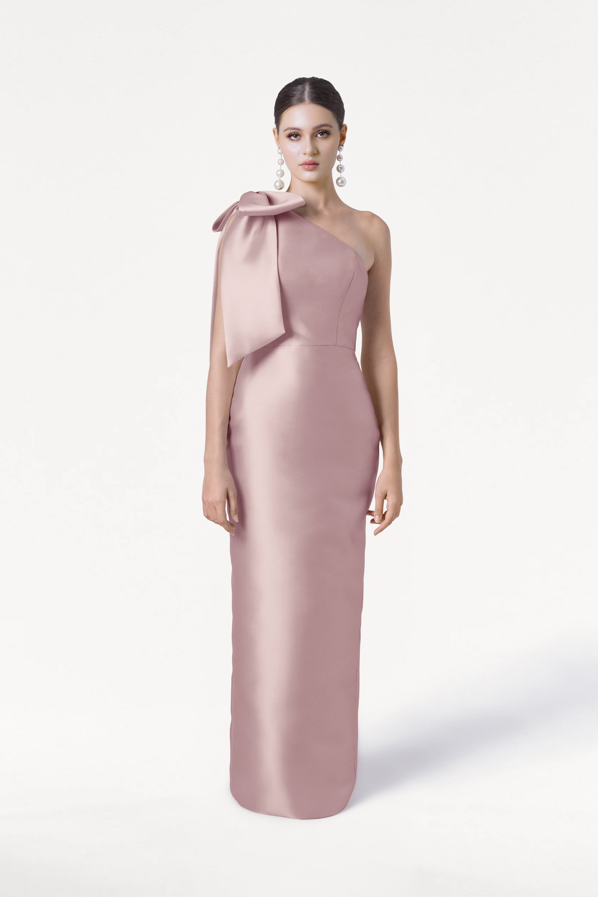 A woman with dark hair pulled back and large dangling earrings models the TH&TH Ritz Bridesmaid Dress in Smoked Blush Satin Twill, a long one-shoulder gown with a statement bow, against a plain white background.