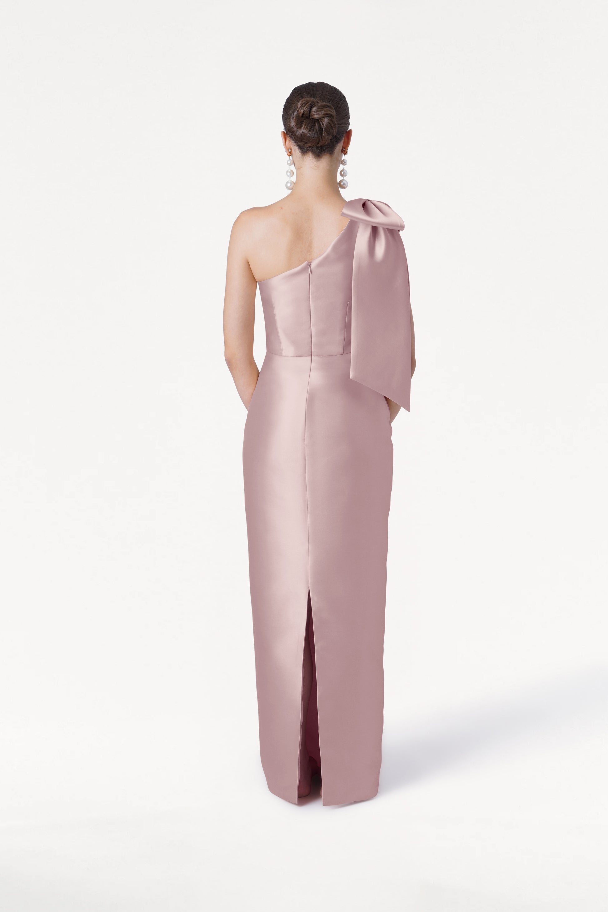 A woman faces away, wearing the TH&TH Ritz Bridesmaid Dress in Smoked Blush Satin Twill—a light pink one-shoulder gown with a statement bow and back slit. Her hair is in a low bun, with large earrings, set against a plain white background.