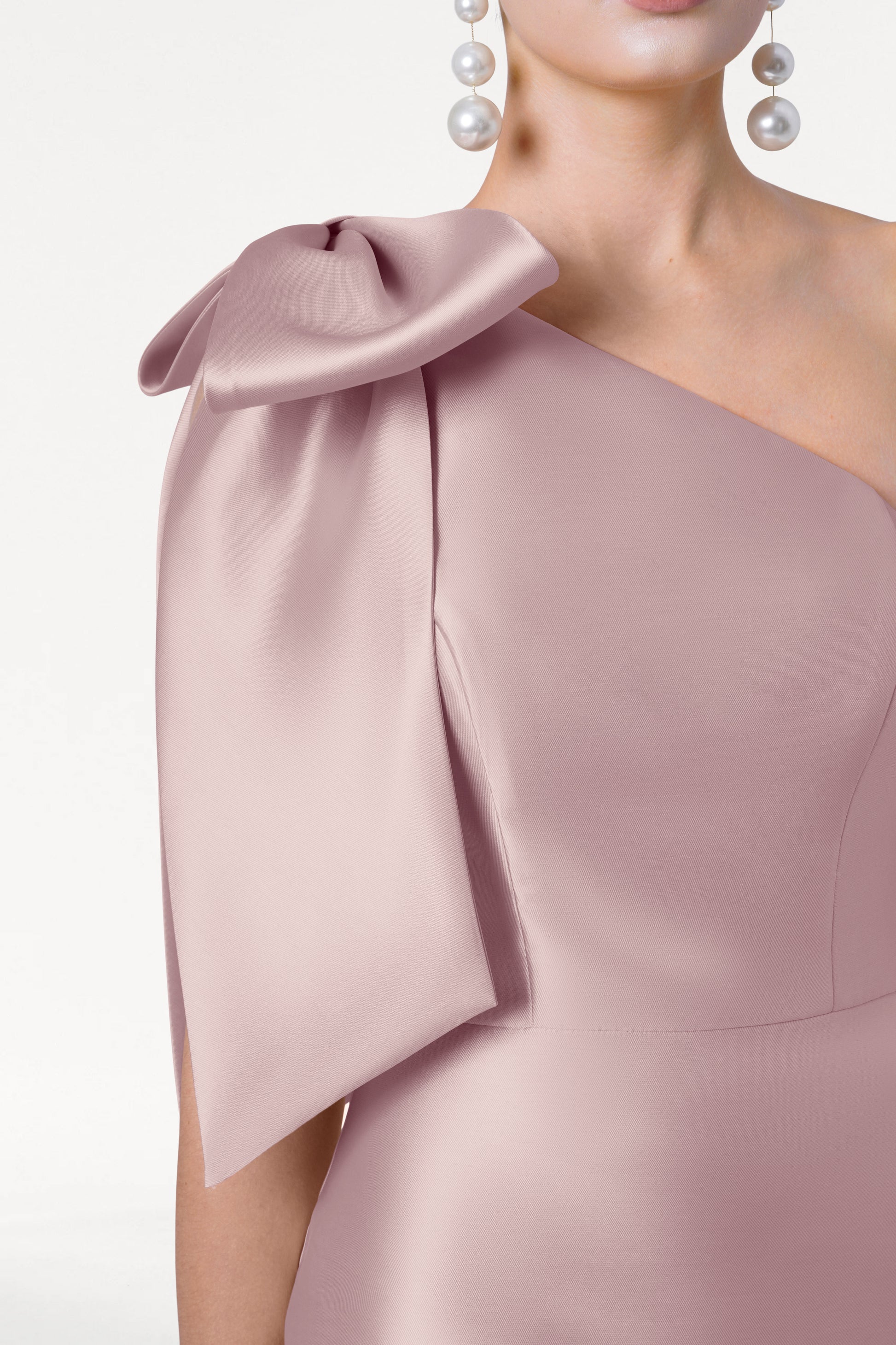 A close-up of a woman in the TH&TH Ritz Bridesmaid Dress in Smoked Blush Satin Twill, with a statement one-shoulder bow and pearl drop earrings, against a plain white background. Her face is partially out of frame.