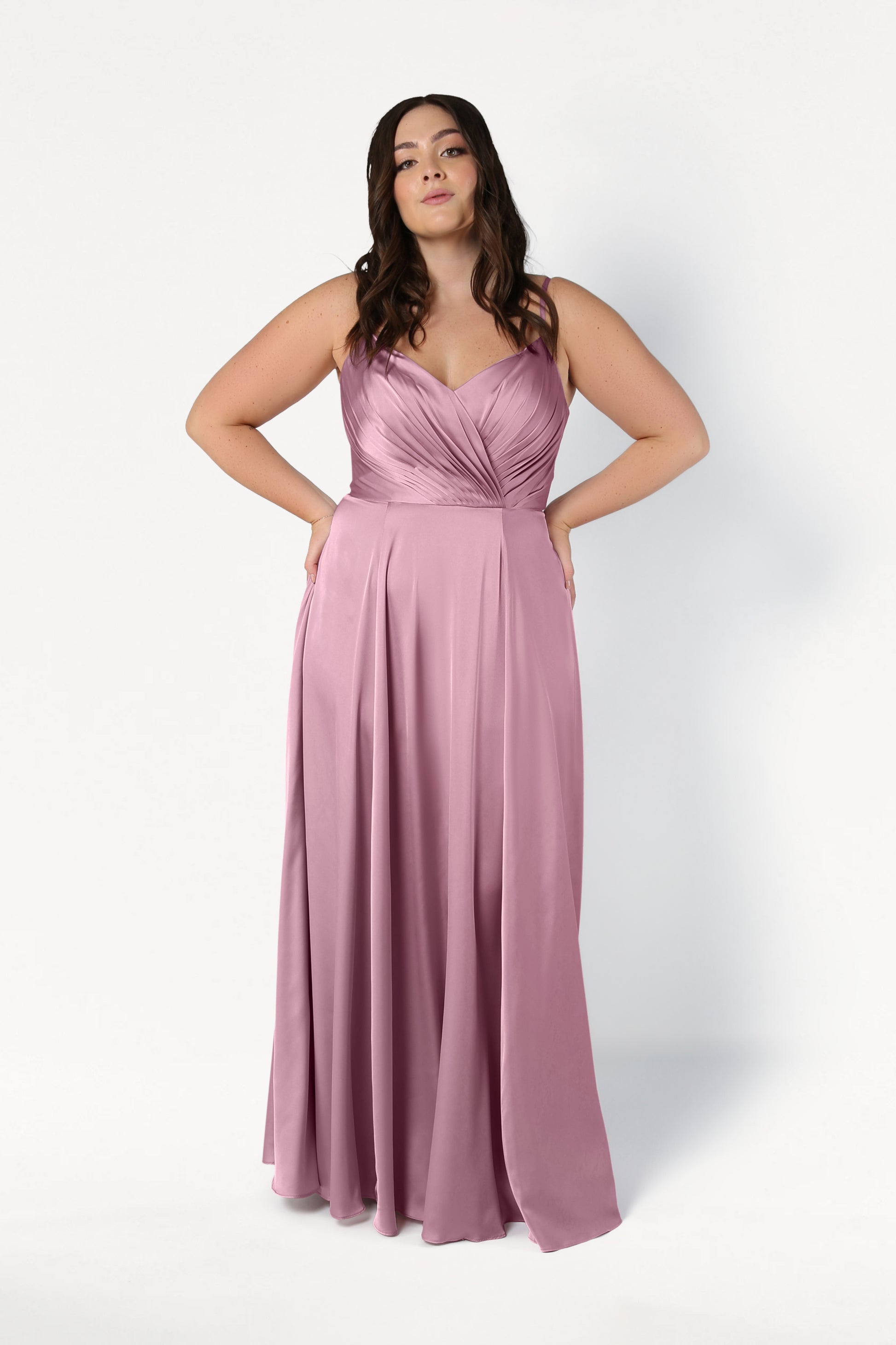 A woman models the TH&TH Isla Bridesmaid Dress in rose pink satin, featuring a sleeveless, floor-length design with a pleated bodice and flowing skirt, standing against a plain white background with hands on her hips.