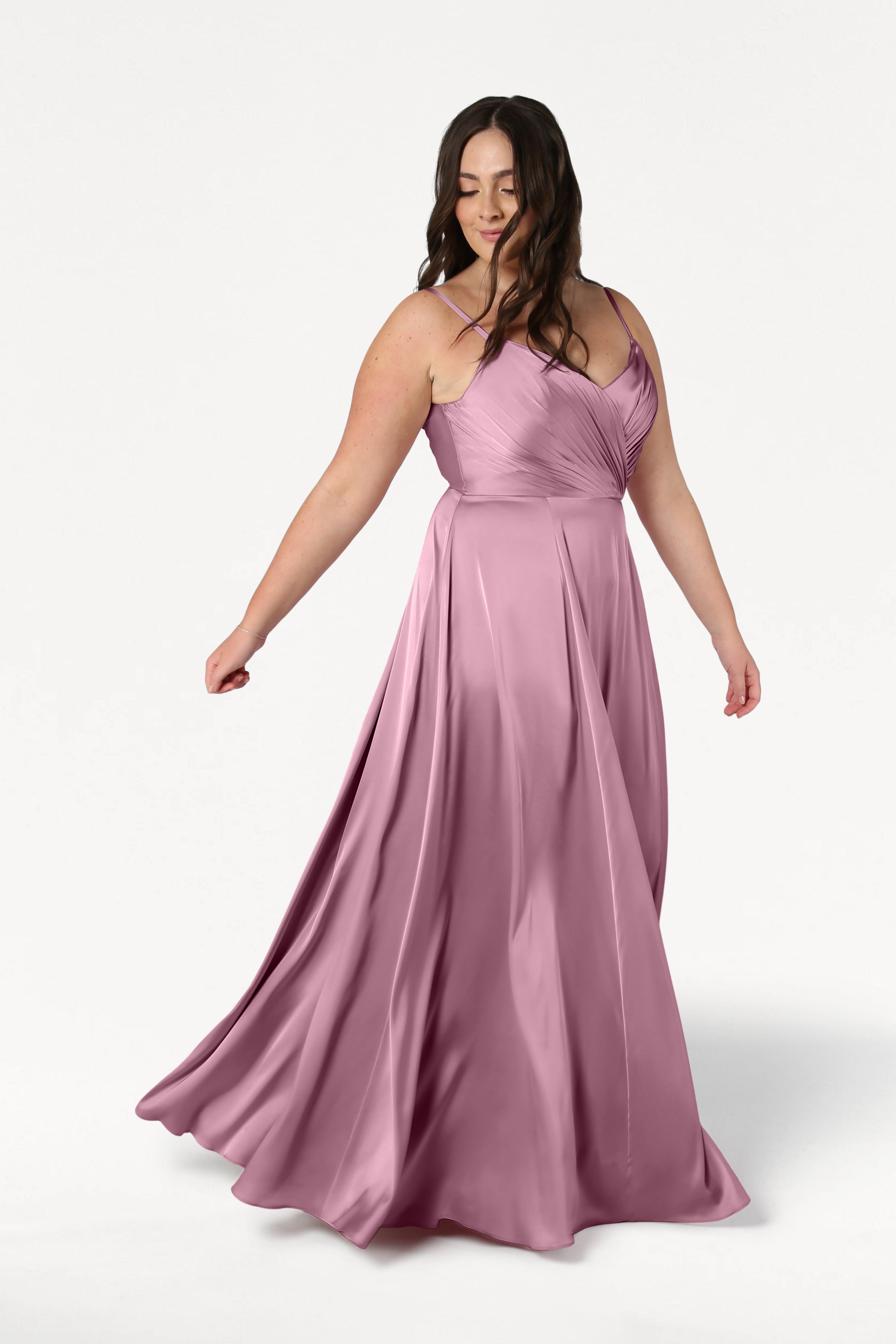 A woman with long brown hair wears the TH&TH Isla Bridesmaid Dress in rose pink satin, featuring a flowing, sleeveless design with a pleated bodice, as she stands against a white background and gently holds the swaying gown.