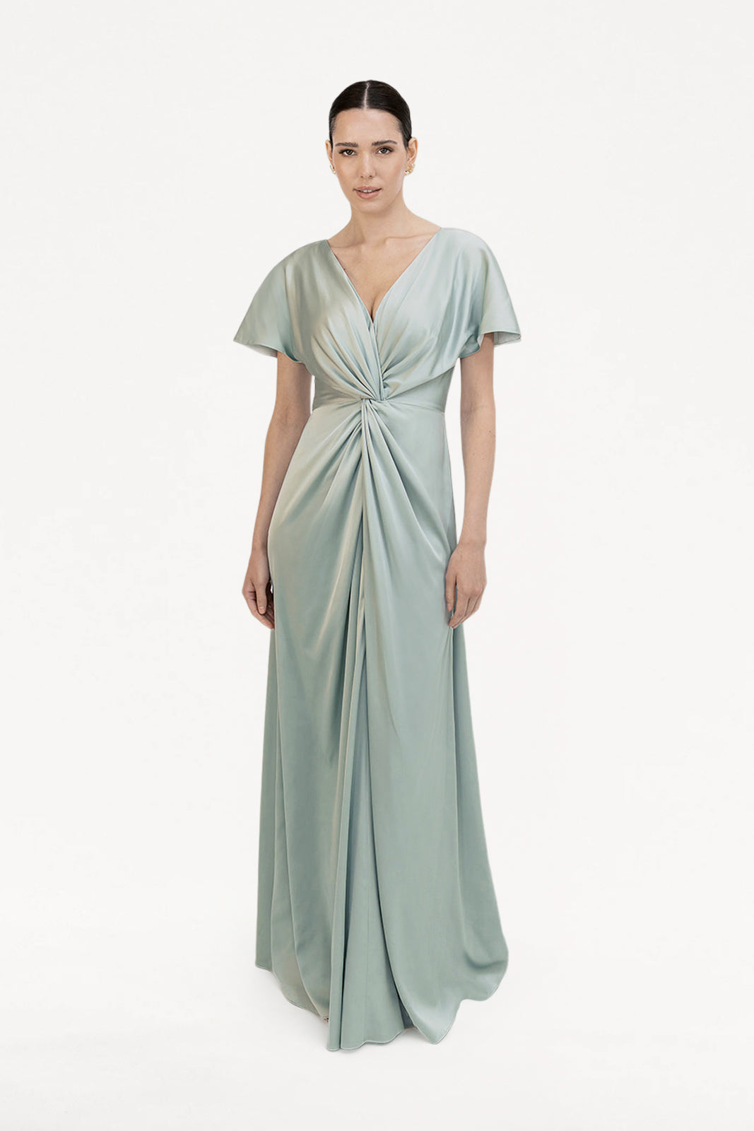 A woman with dark hair in a sleek, low bun models the TH&TH Camilla Bridesmaid Dress in Dusty Eucalyptus Satin, a size-inclusive gown featuring a twist-knot bodice, short sleeves, and gathered waist against a white background.