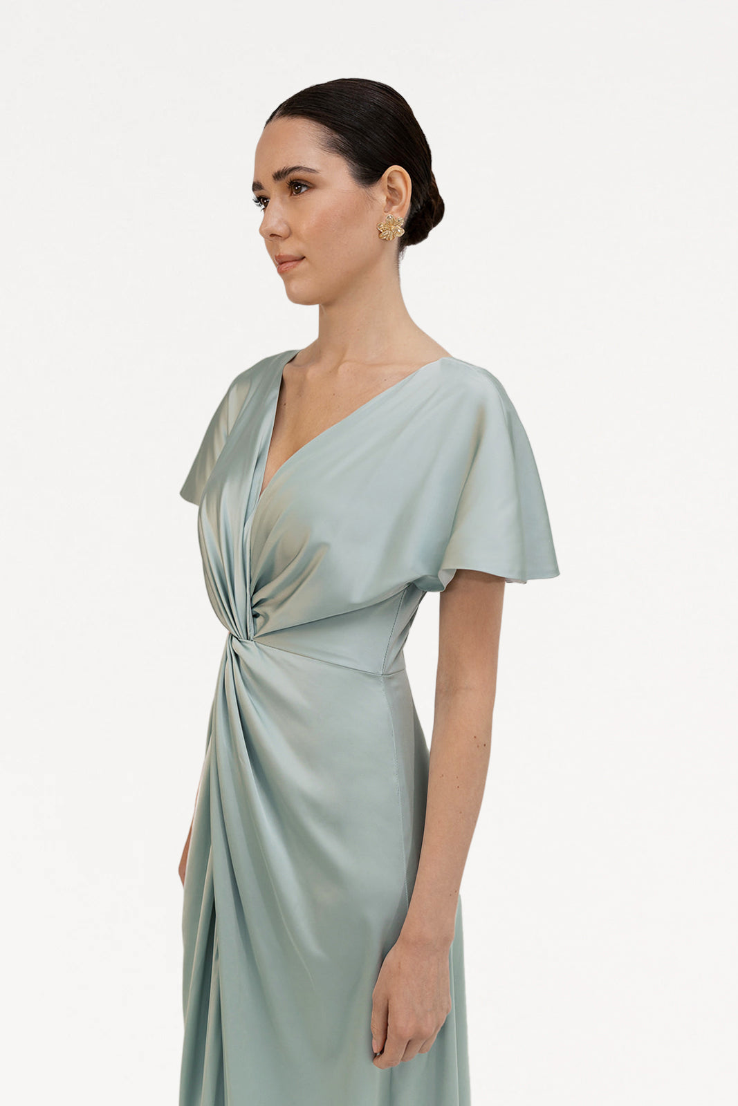 A woman with dark hair in a low bun wears the TH&TH Camilla Bridesmaid Dress in Dusty Eucalyptus Satin—this size-inclusive gown features short sleeves and a twist-knot bodice, shown against a plain white background.