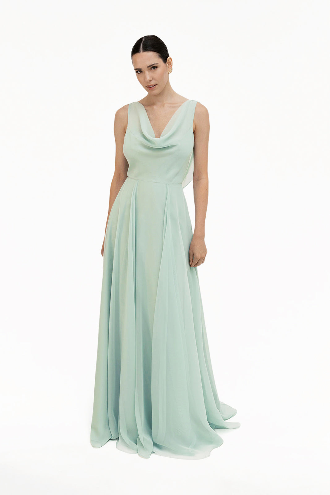 A woman poses against a plain white background in the Athena Bridesmaid Dress in Dusty Eucalyptus by TH&TH—a sleeveless, floor-length chiffon gown with a draped cowl neck. Her dark hair is pulled back.