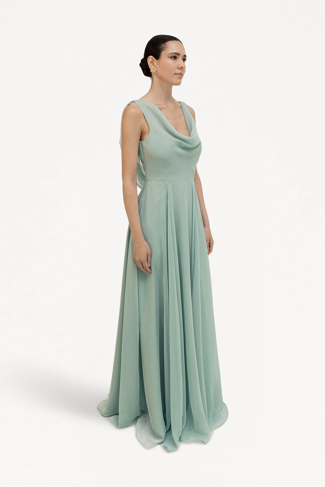 A woman with dark hair pulled back wears the TH&TH Athena Bridesmaid Dress in Dusty Eucalyptus, featuring a long, flowing chiffon silhouette and draped cowl neck, standing against a plain white background looking slightly to the side.