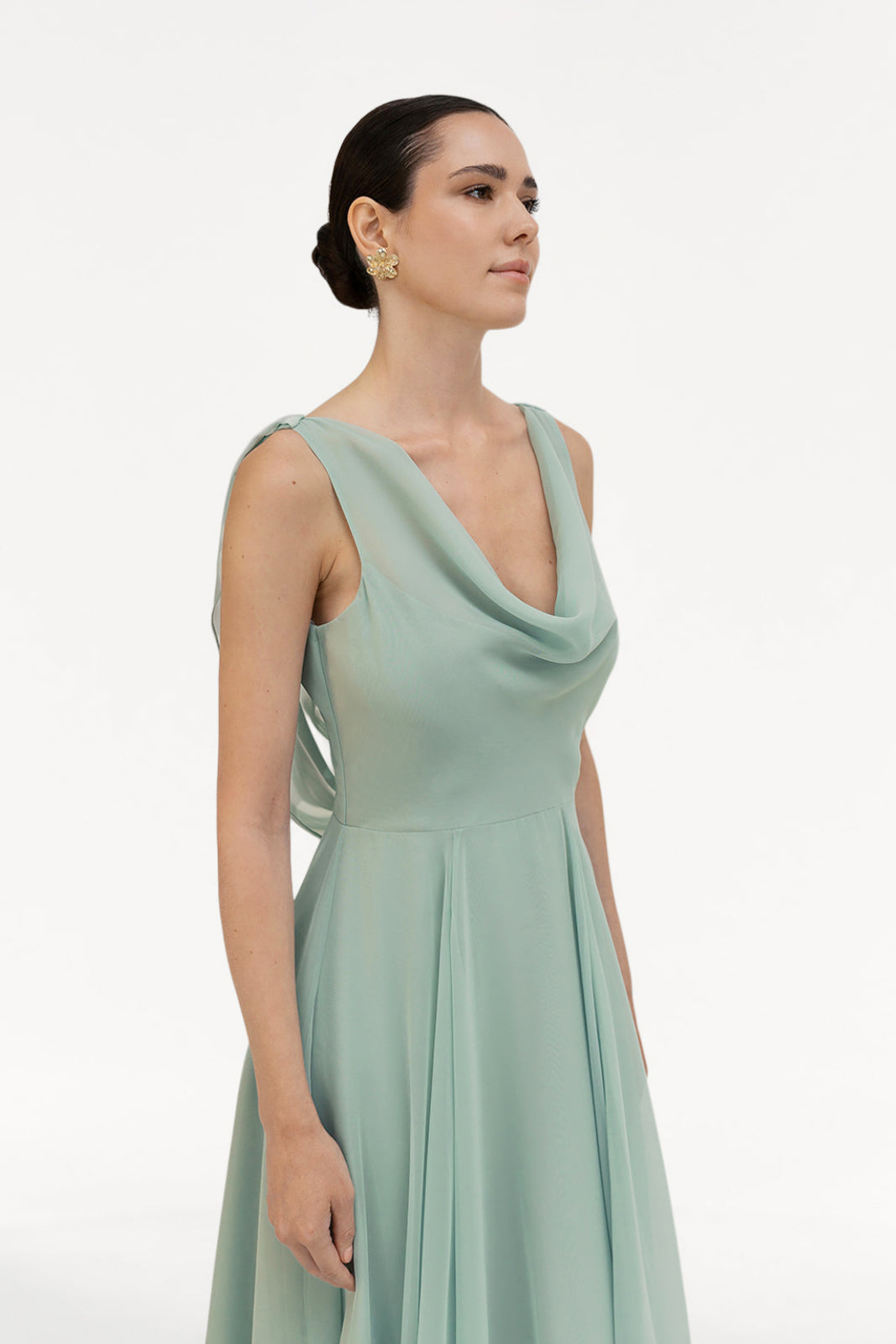 A woman with dark hair in a low bun wears the TH&TH Athena Bridesmaid Dress in Dusty Eucalyptus, a sleeveless chiffon gown with a cowl neck, paired with gold earrings, standing against a plain white background.