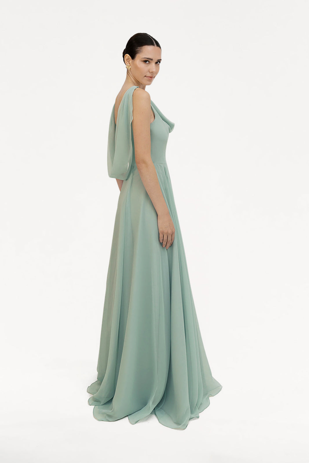 A woman stands in profile wearing the TH&TH Athena Bridesmaid Dress in Dusty Eucalyptus, featuring flowing chiffon with draped fabric over one shoulder, set against a plain white background.