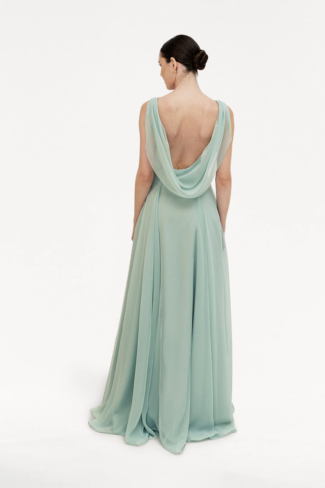 A woman faces away, wearing the TH&TH Athena Bridesmaid Dress in Dusty Eucalyptus—a flowing, floor-length chiffon gown with a draped cowl neck and low back. Her hair is neatly styled in a bun, set against a plain white background.
