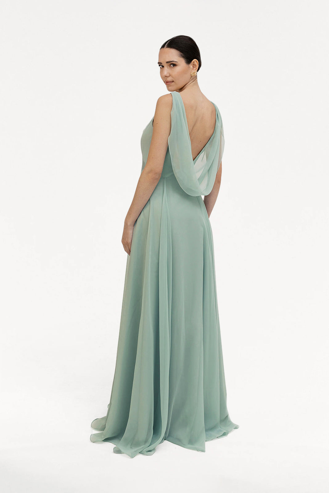 A woman faces backward, looking over her shoulder in the TH&TH Athena Bridesmaid Dress in Dusty Eucalyptus, featuring a flowing chiffon fabric with a cowl neck and draped back against a plain white background.