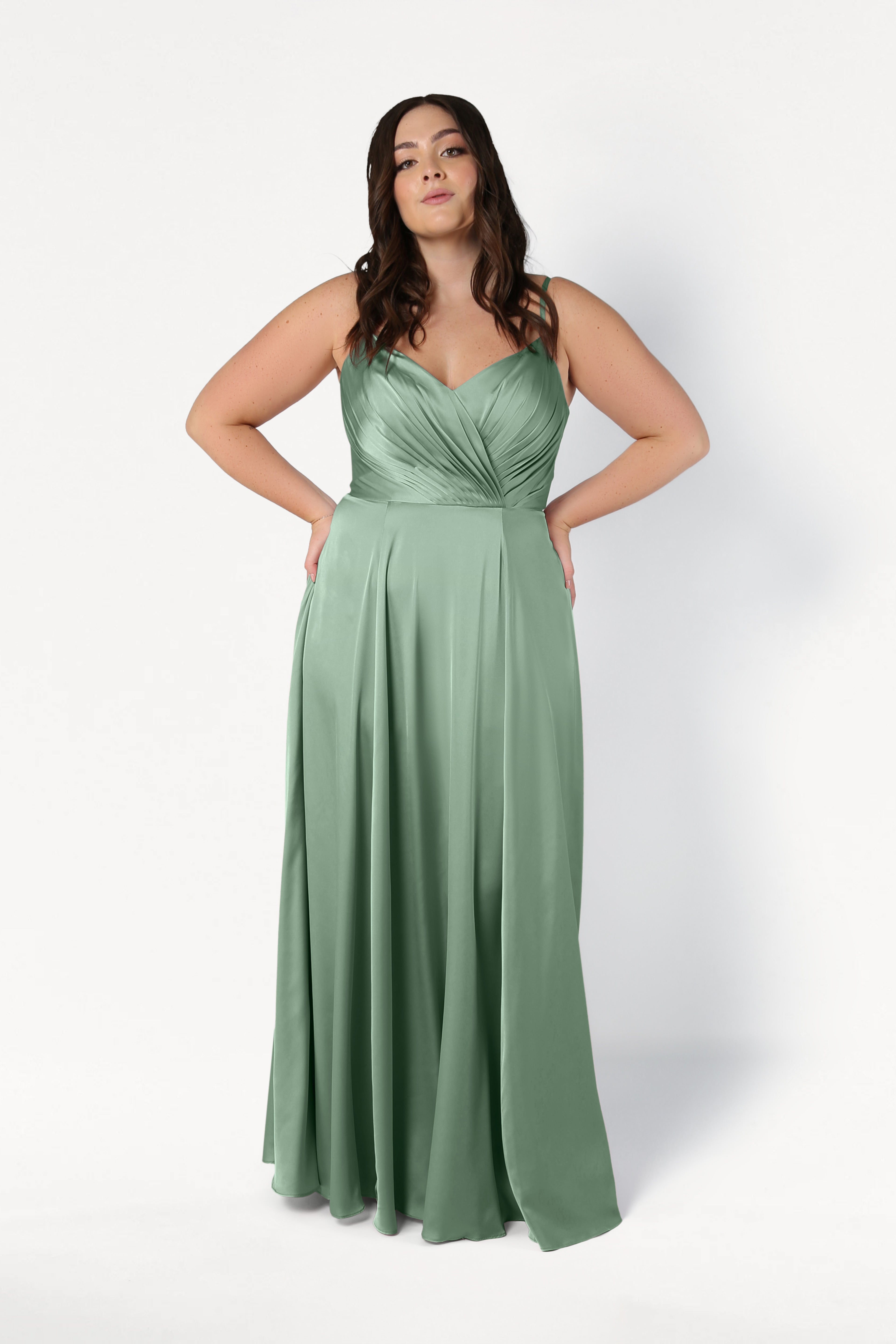 A woman with long, wavy dark hair stands against a white background in the TH&TH Isla Bridesmaid Dress—a floor-length sage green satin dress with a pleated bodice and skirt—her hands resting lightly on her hips.